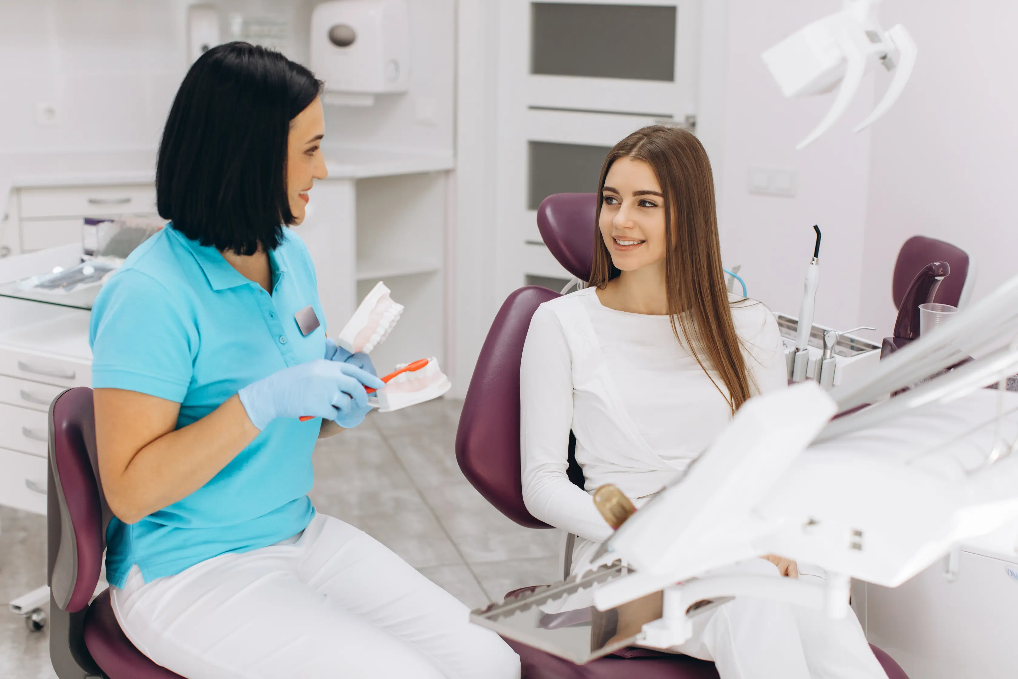 Female dentist in blue scrubs discussing oral care and treatment options with a female patient seated in a dental chair during a smile consultation