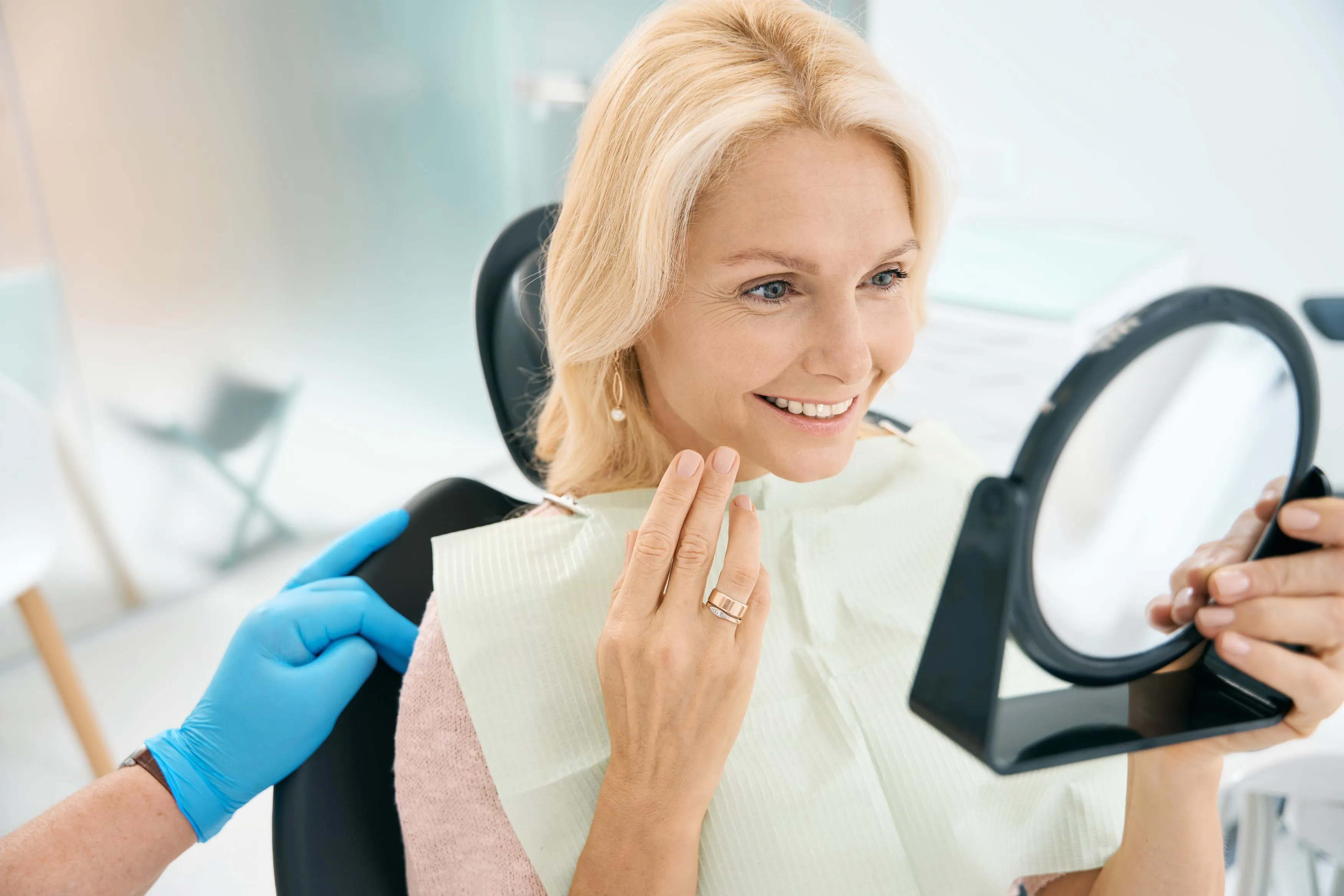 Smiling female patient sitting in a dental chair holding a mirror and admiring her bright new smile after completing a cosmetic whitening treatment