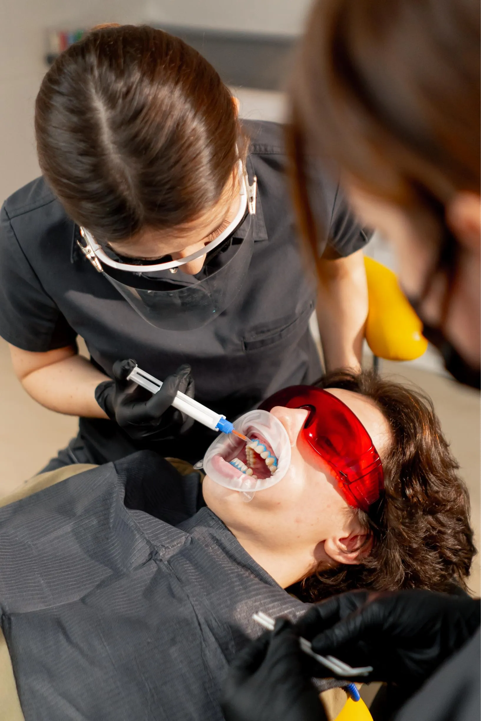 Female dentist wearing protective eyewear using a laser whitening device on a patient reclined in the chair during a professional cosmetic dental procedure