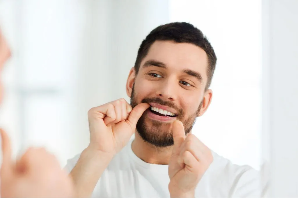 Young man smiling while flossing his teeth in front of a mirror emphasizing healthy dental habits and at-home oral hygiene maintenance