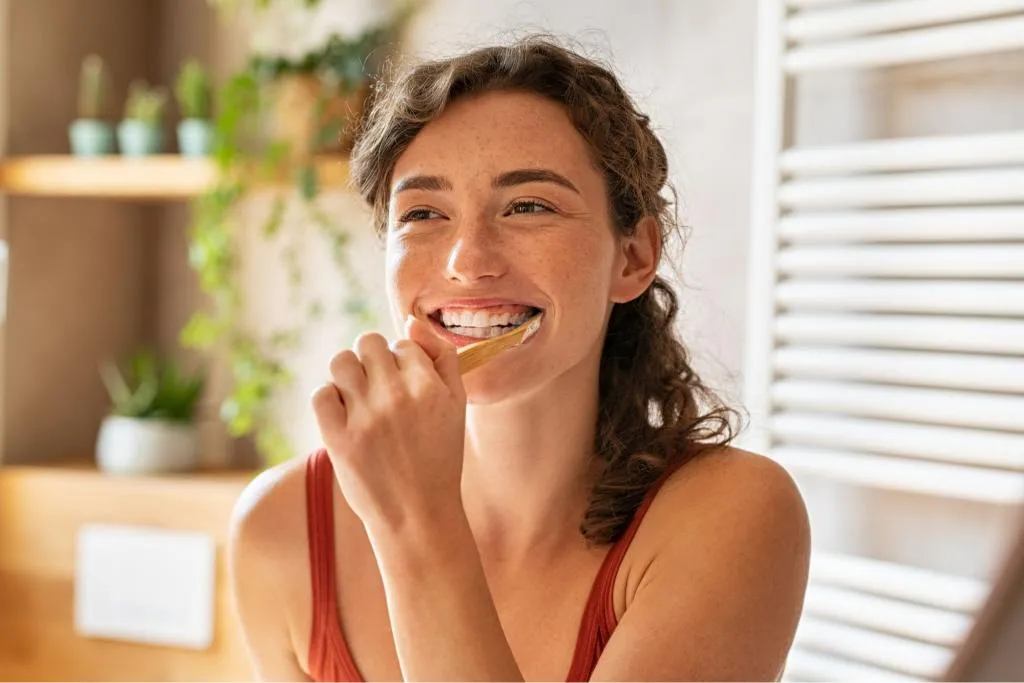 Smiling woman brushing her teeth in a bright bathroom with natural light representing good oral hygiene habits and a healthy daily dental care routine