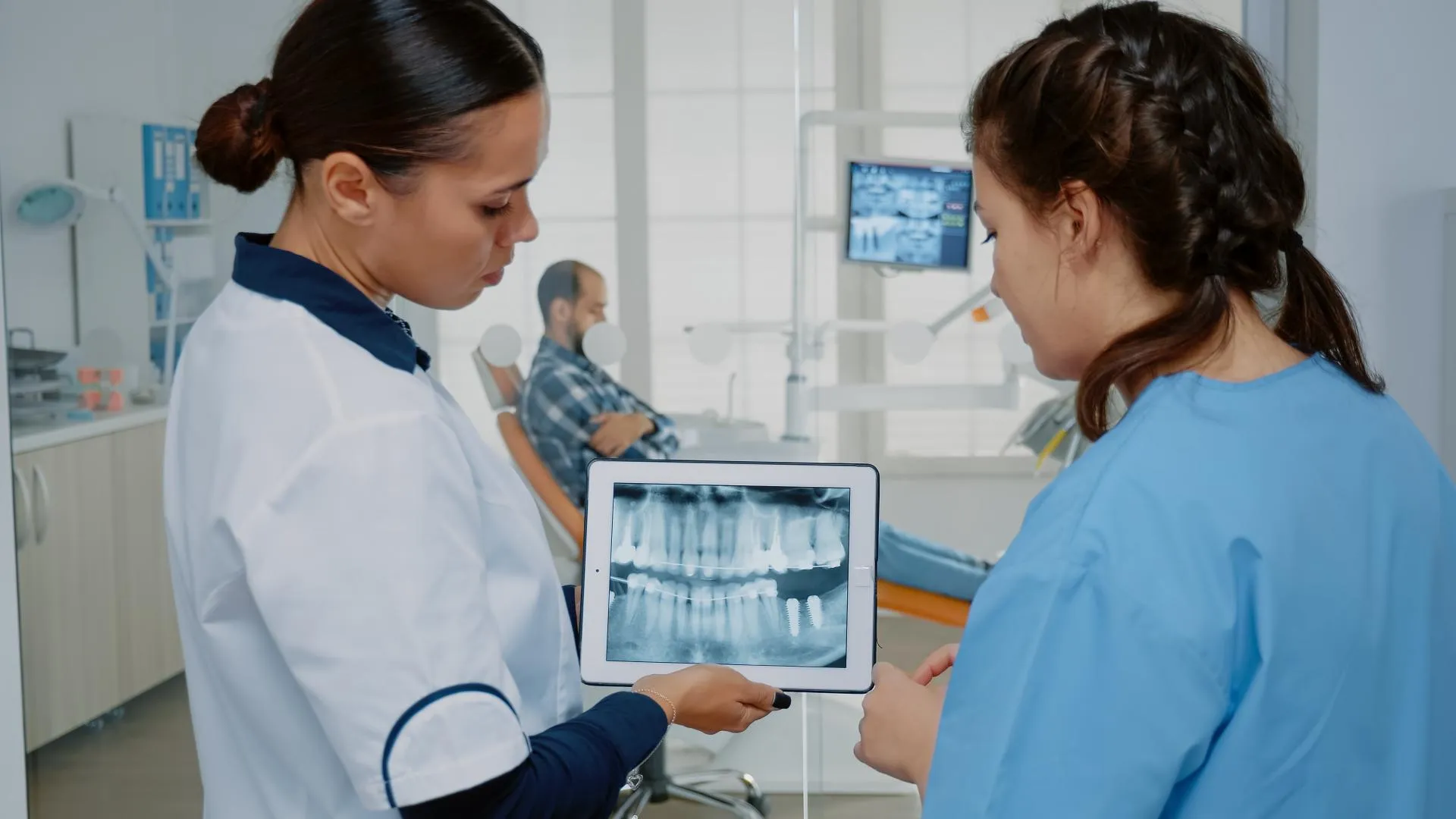 Two dentists in clinical uniforms reviewing a patient’s dental X-ray on a tablet in a bright, modern dental office while planning a treatment procedure