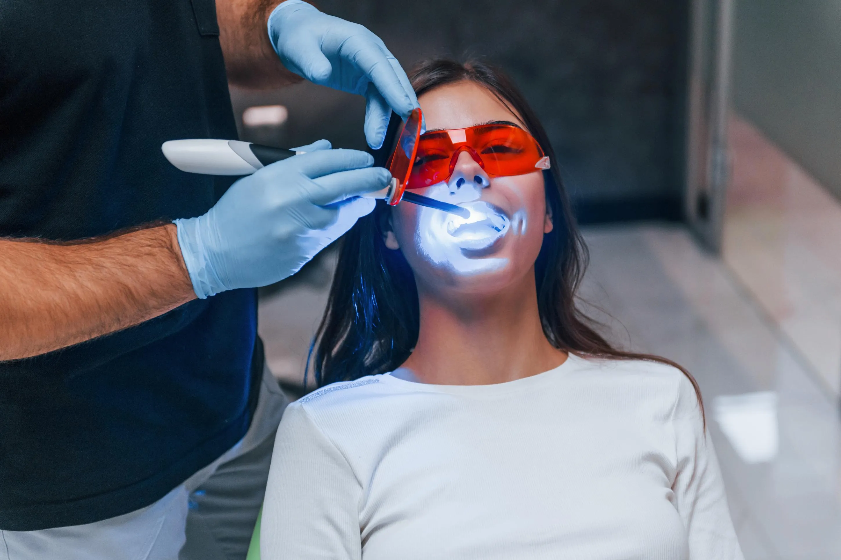 Female patient wearing red protective glasses receiving laser teeth whitening treatment performed by a dental professional to enhance smile brightness and remove stains