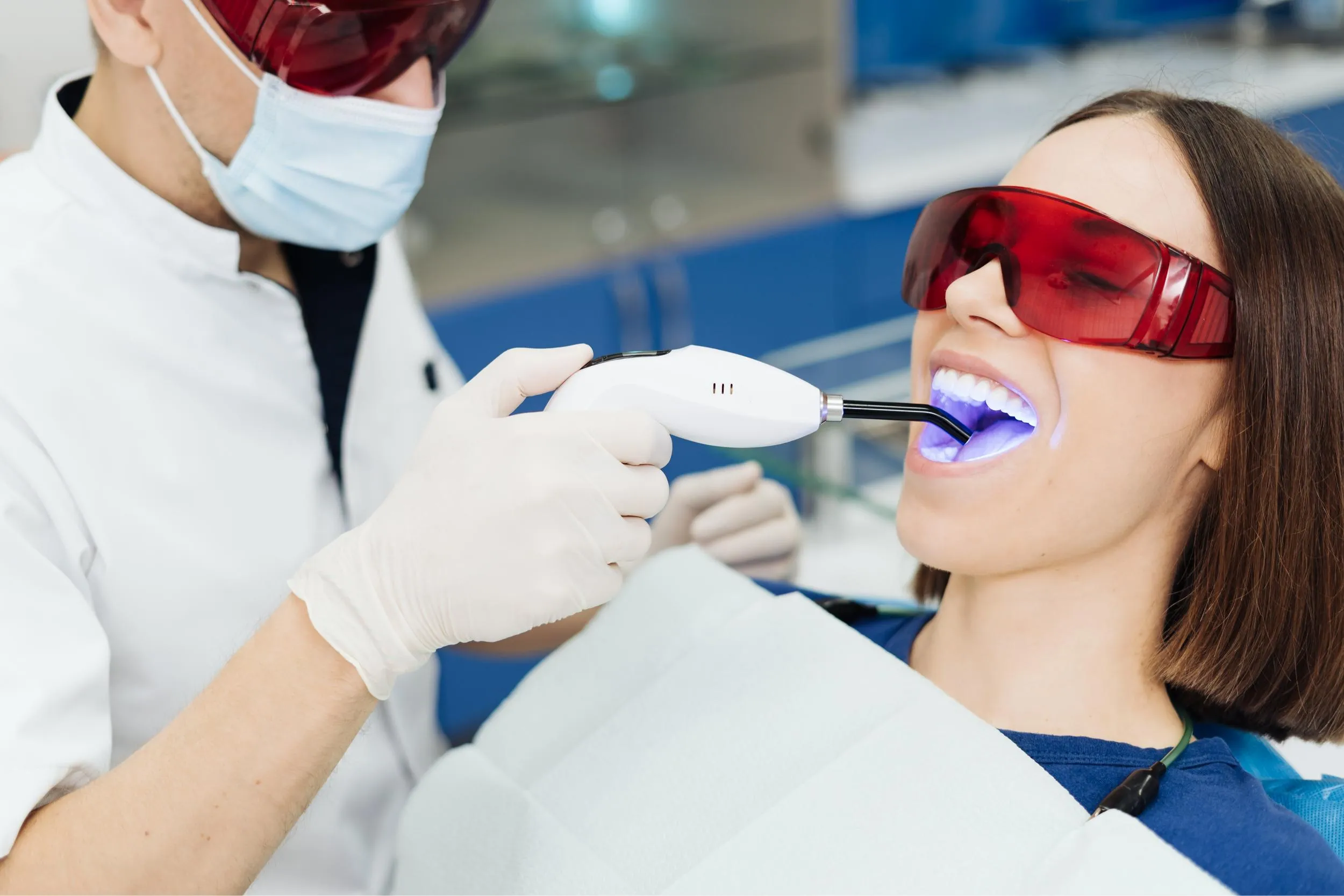 Dentist in white coat performing a laser teeth whitening procedure on a female patient wearing red safety glasses in a high-tech cosmetic dental clinic