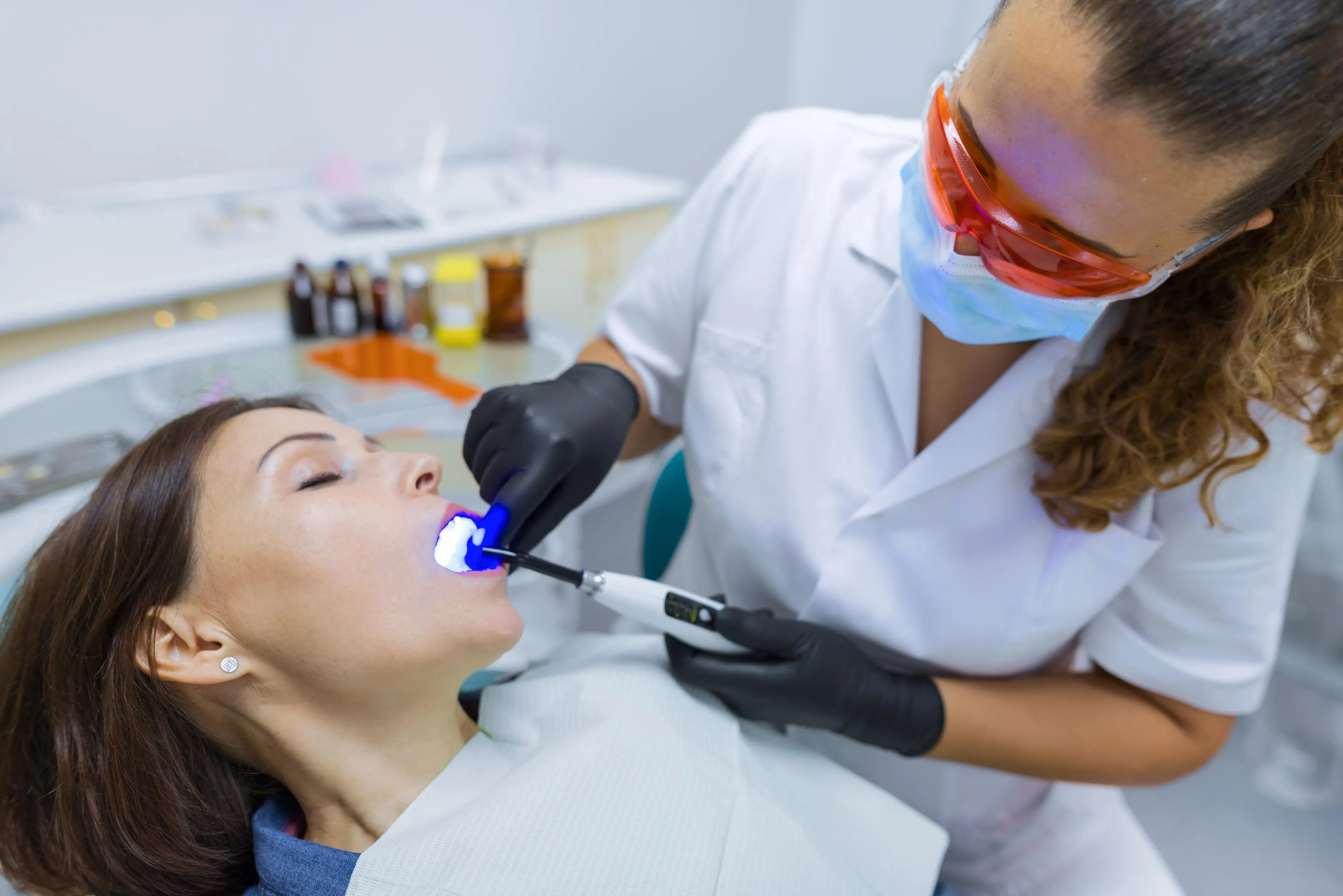 Dentist wearing protective glasses using a blue LED light to perform professional teeth whitening on a female patient in a modern dental office