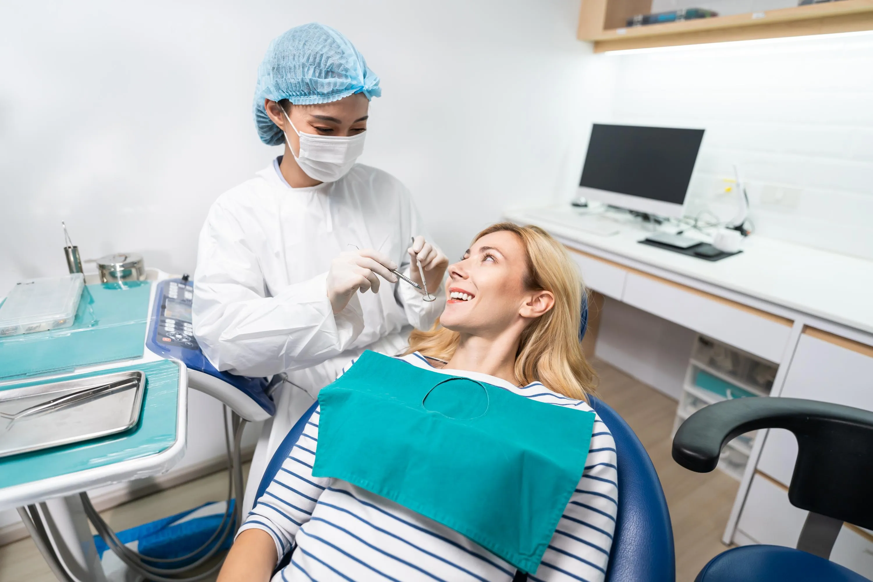 Dentist in surgical mask and protective eyewear examining a female patient’s teeth using a dental mirror during a routine oral health assessment