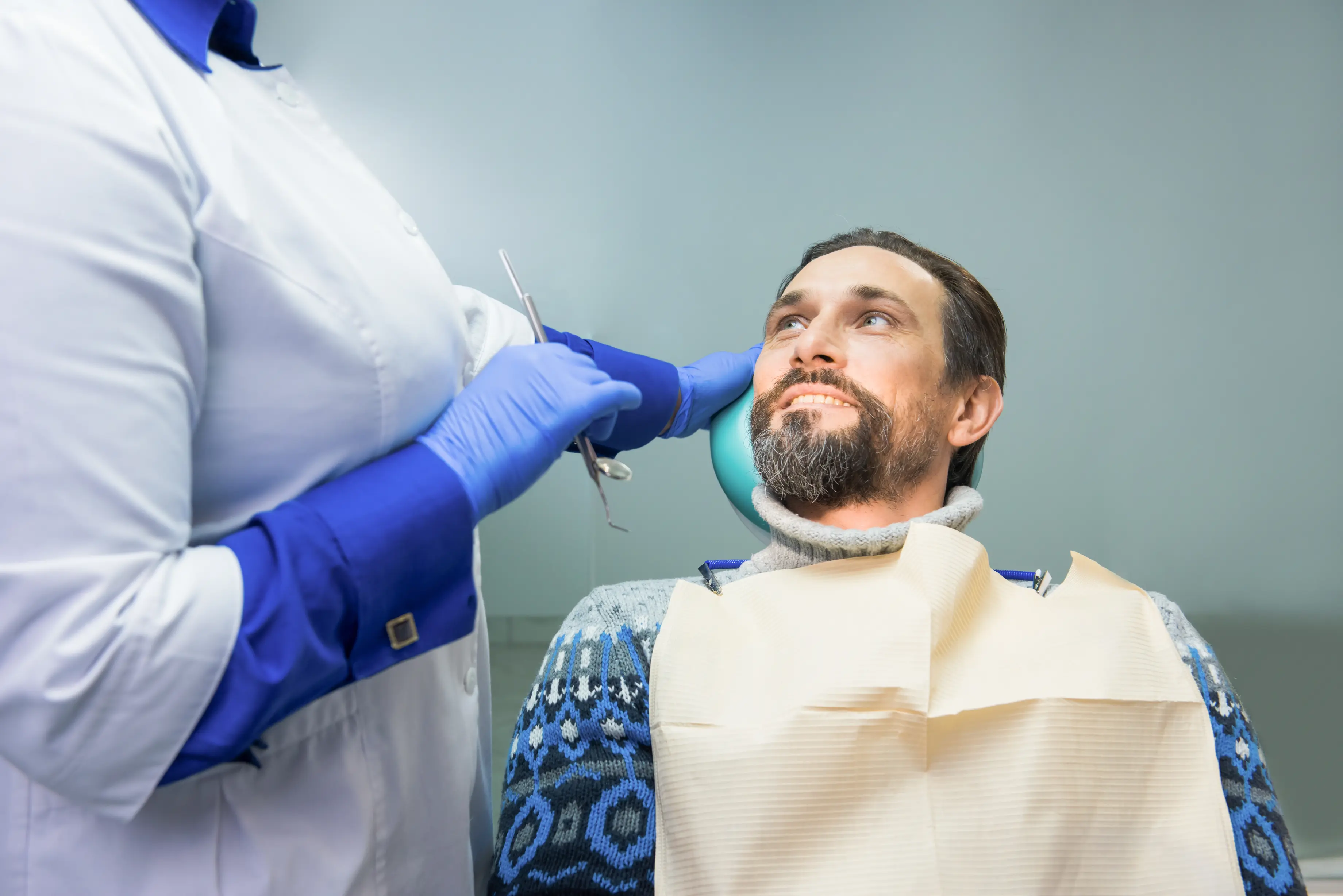 Dentist in white coat and blue gloves examining a male patient sitting in a dental chair during a routine oral health checkup emphasizing preventive care and comfort