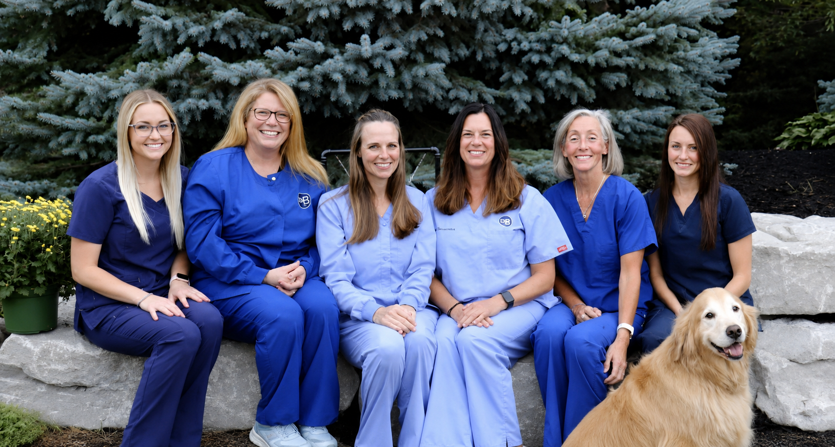 Group of six dental team members sitting outdoors in front of green trees, all dressed in various shades of blue scrubs and smiling warmly, accompanied by a friendly golden retriever at their side