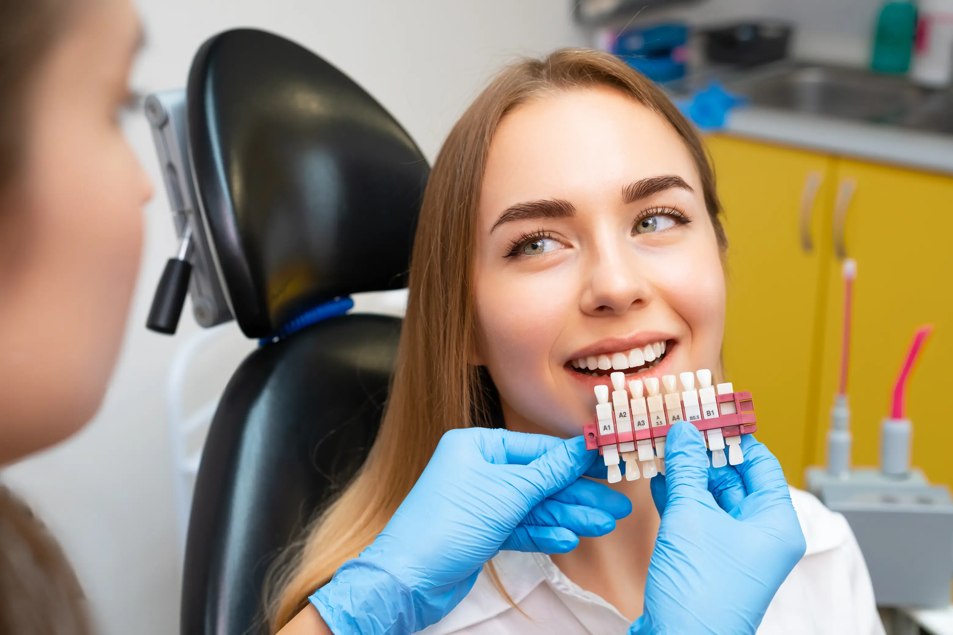 Female dental patient reclined in a treatment chair while a dentist wearing purple gloves holds a tooth shade guide next to her smile to evaluate tooth color for whitening or restoration