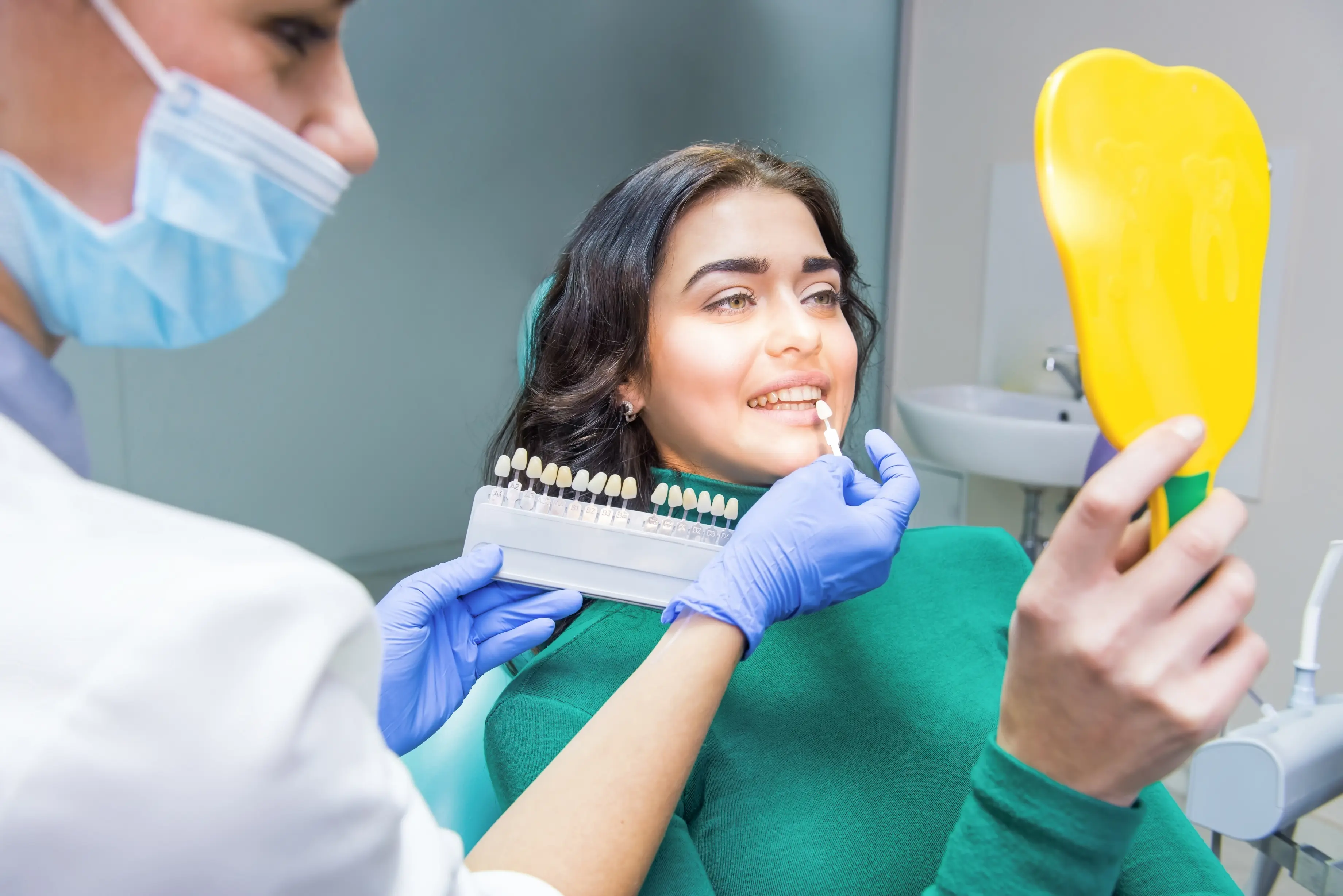 Female dental patient smiling while a dentist in blue gloves holds a tooth shade guide near her mouth, comparing shades for whitening or veneer treatment in a brightly lit dental clinic