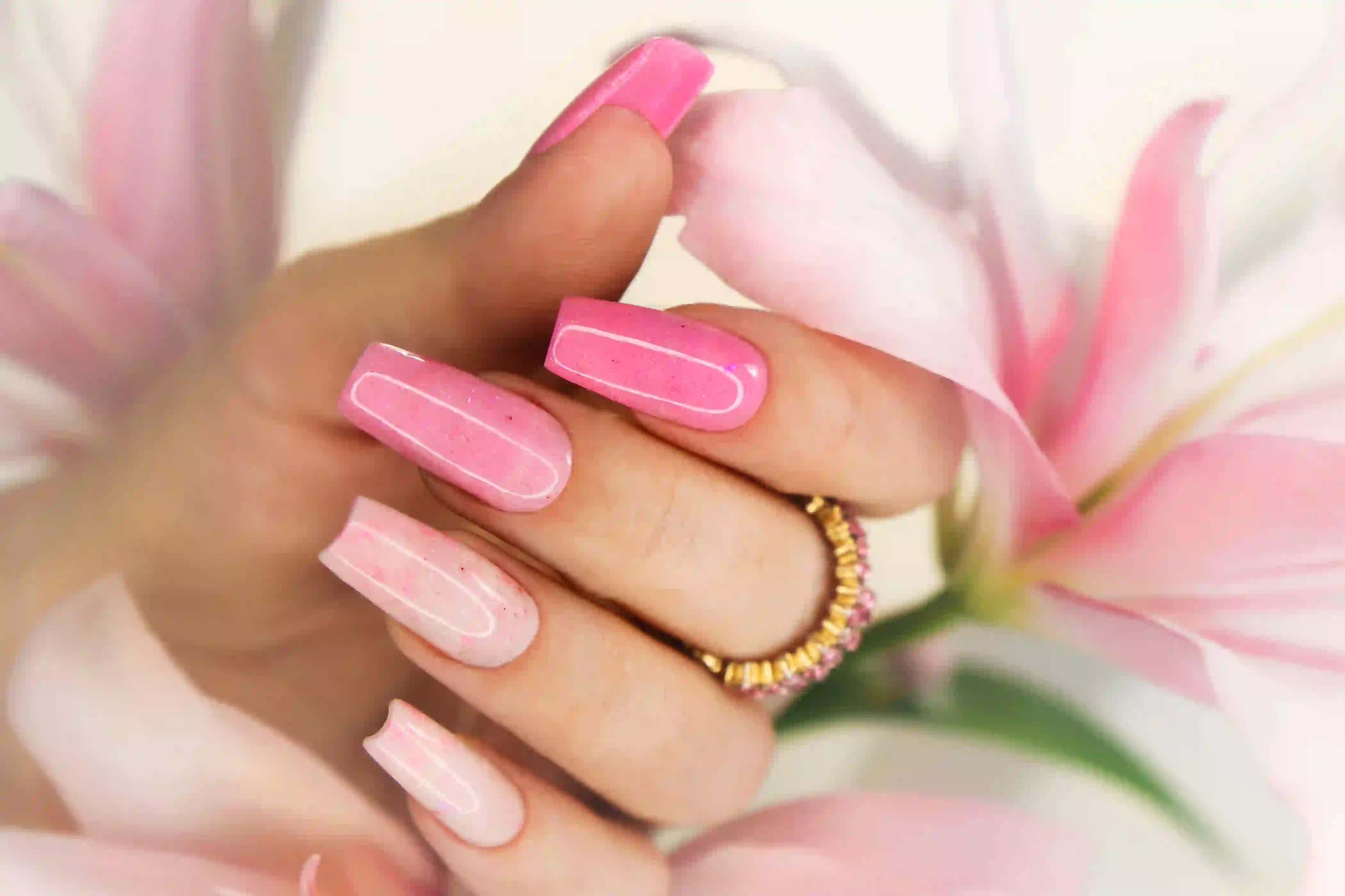 Close-up of a hand with glossy pink ombre nails holding a delicate pink flower, wearing a gold and pink gemstone ring.