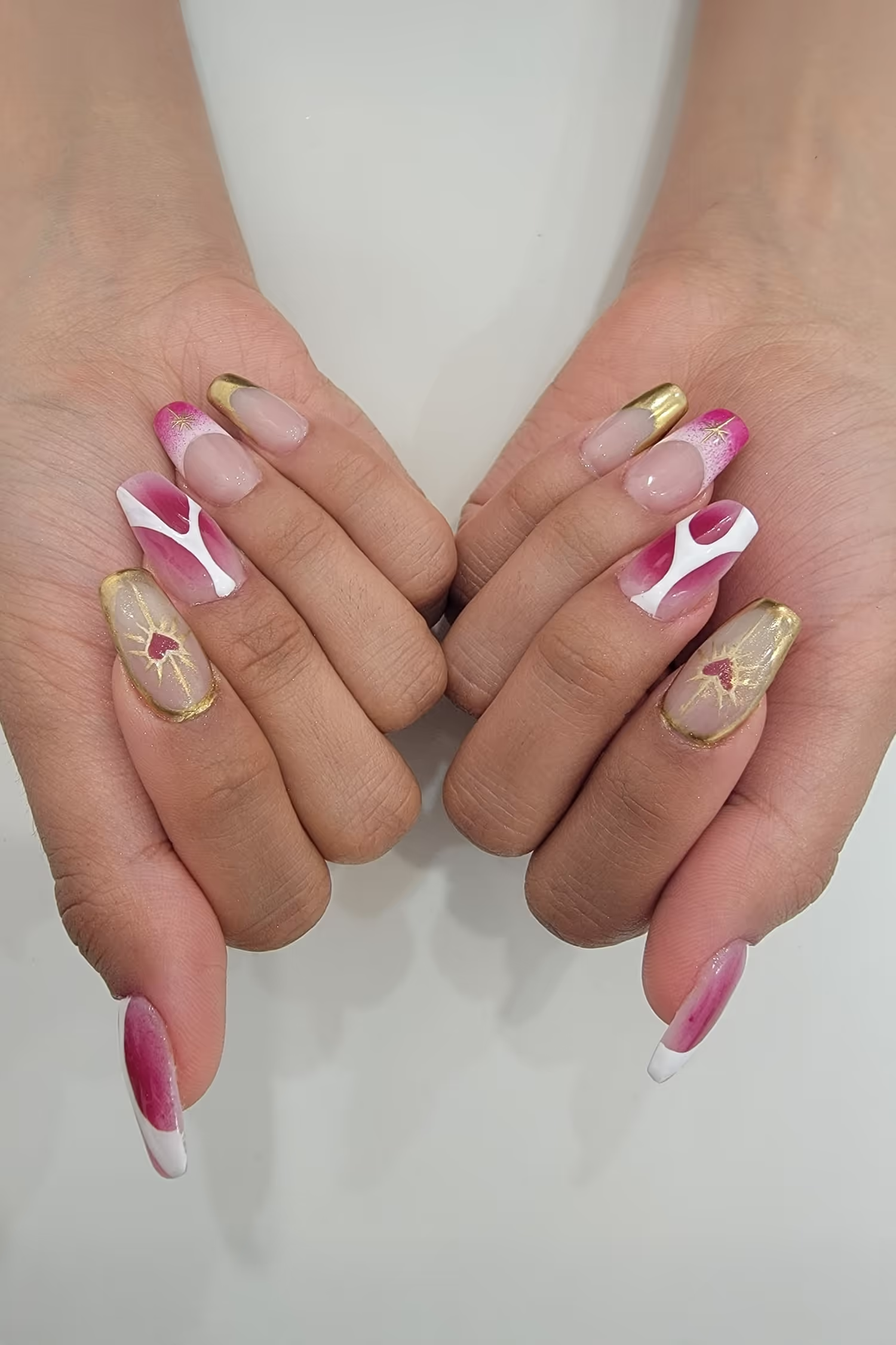 Hands with long nails decorated in pink, white, and gold designs including hearts and abstract patterns.