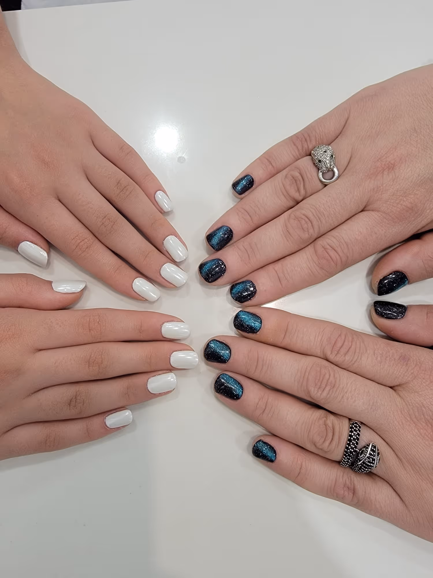 Two pairs of hands with manicured nails facing each other; one with glossy white polish and the other with glittery blue and black polish, wearing silver rings.