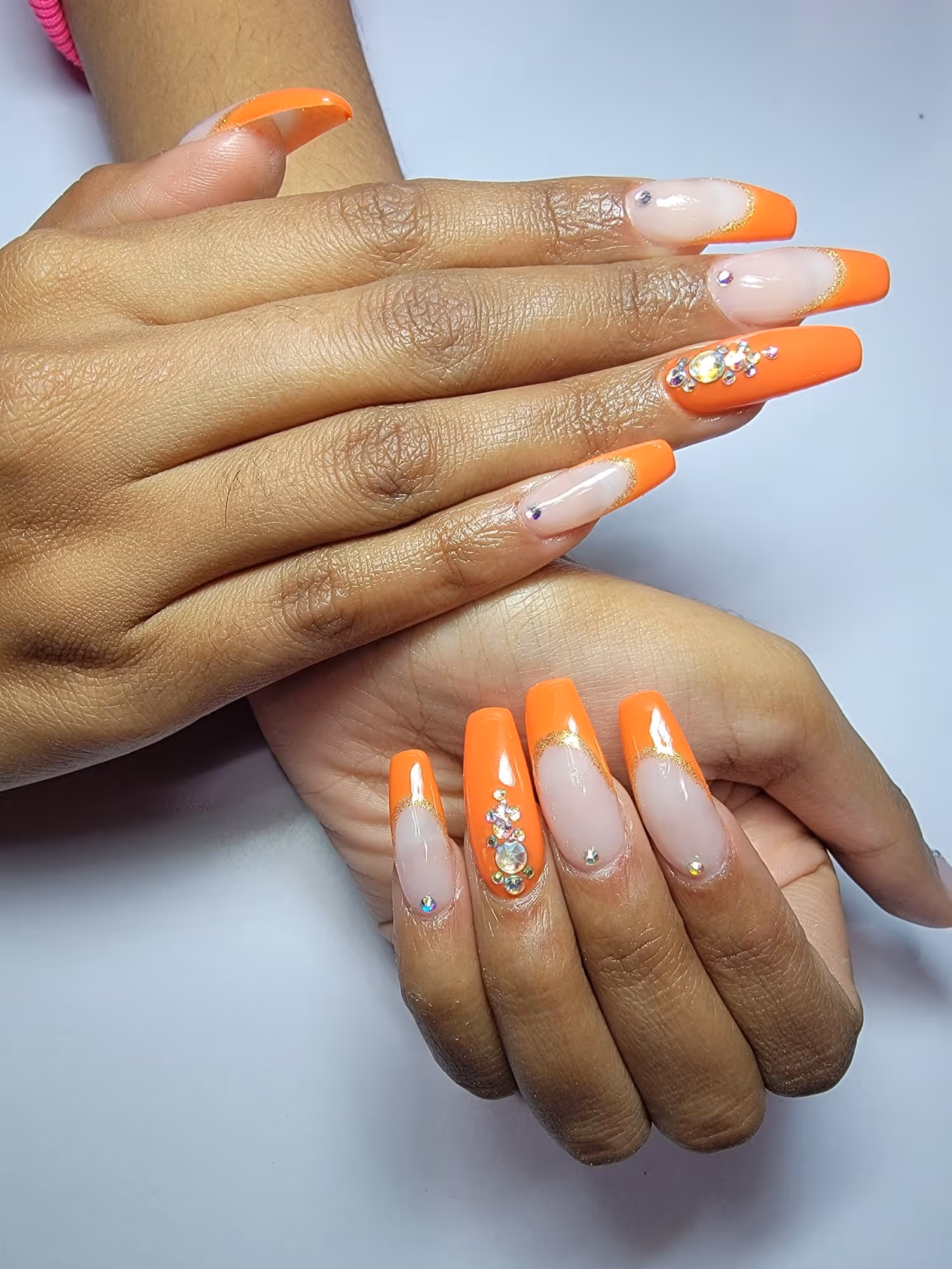 Hands with long coffin-shaped nails featuring a nude base, bright orange tips, gold accents, and rhinestone decorations.