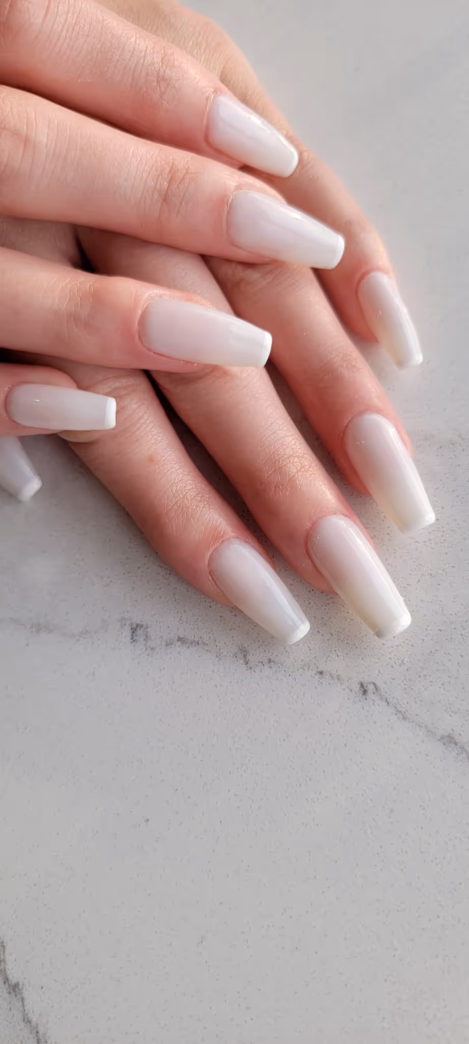 Hands with long, square-shaped glossy pale pink acrylic nails resting on a light gray marble surface.