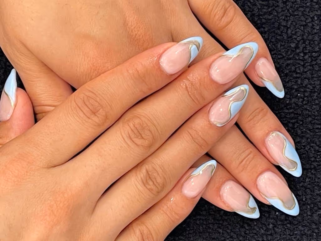 Close-up of hands with almond-shaped nails featuring light blue tips and gold wavy lines over a natural pink base.