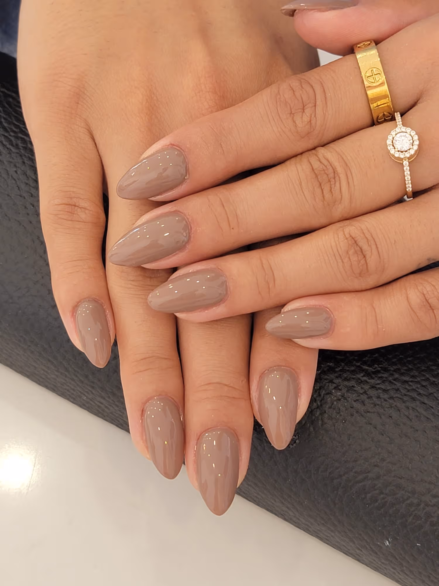 Hands with almond-shaped nails painted in glossy taupe polish and two gold rings, one with a diamond halo setting.
