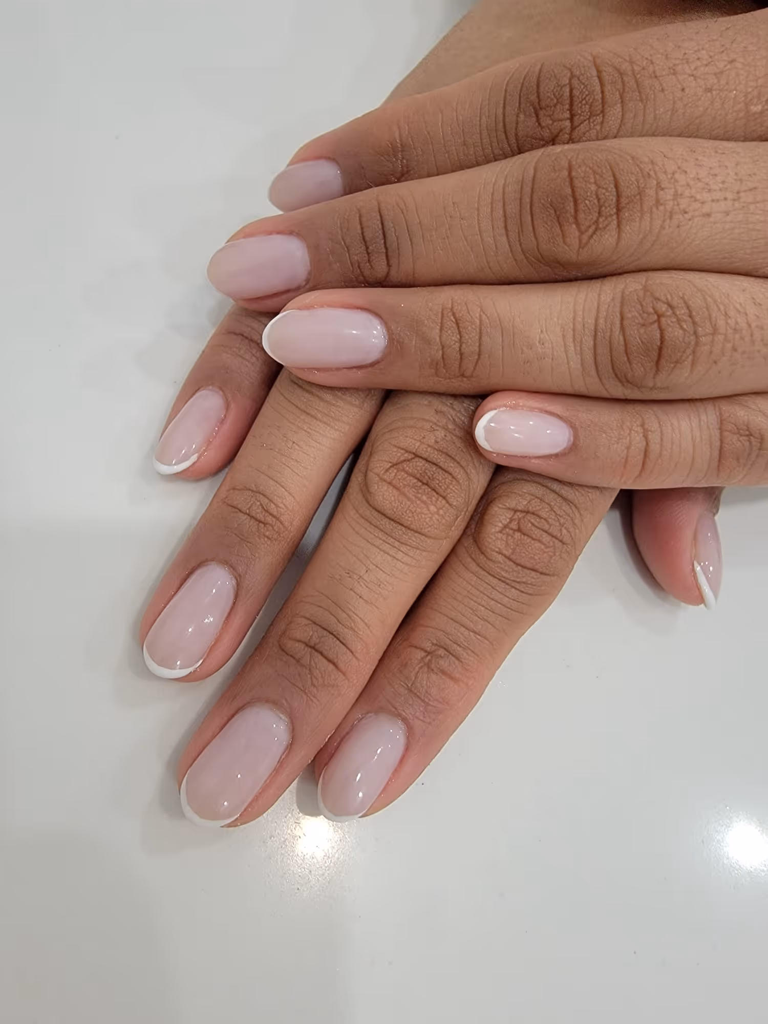 Two hands with light pink French manicure featuring white tips on oval-shaped nails resting on a white surface.