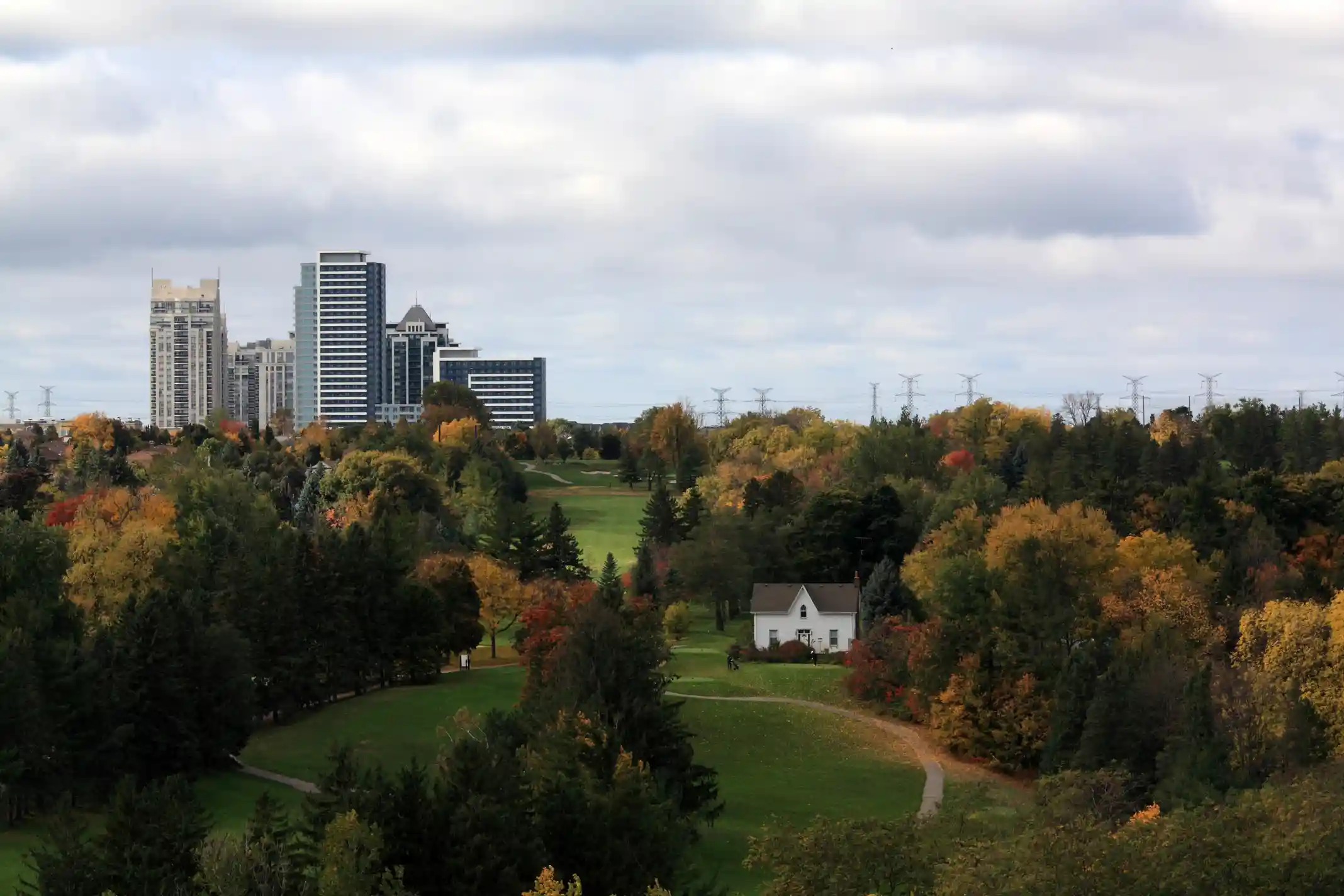 Autumn landscape with colorful trees, a green golf course, a small white house, and a city skyline in the background under a cloudy sky.