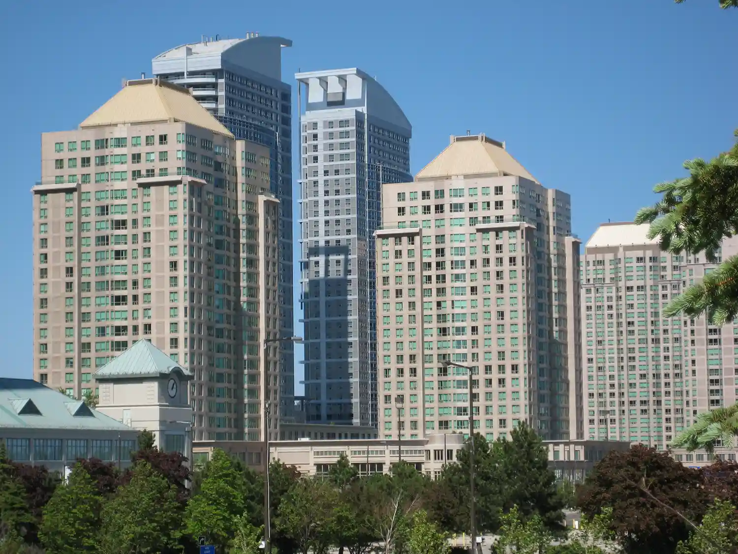 Cluster of modern high-rise buildings with light tan facades and green windows under a clear blue sky, with trees in the foreground.