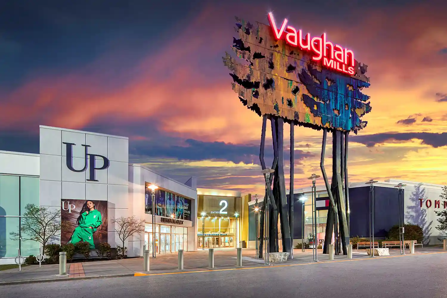 Sunset view of Vaughan Mills shopping mall entrance with a large artistic sign reading 'Vaughan Mills' on tall black supports.
