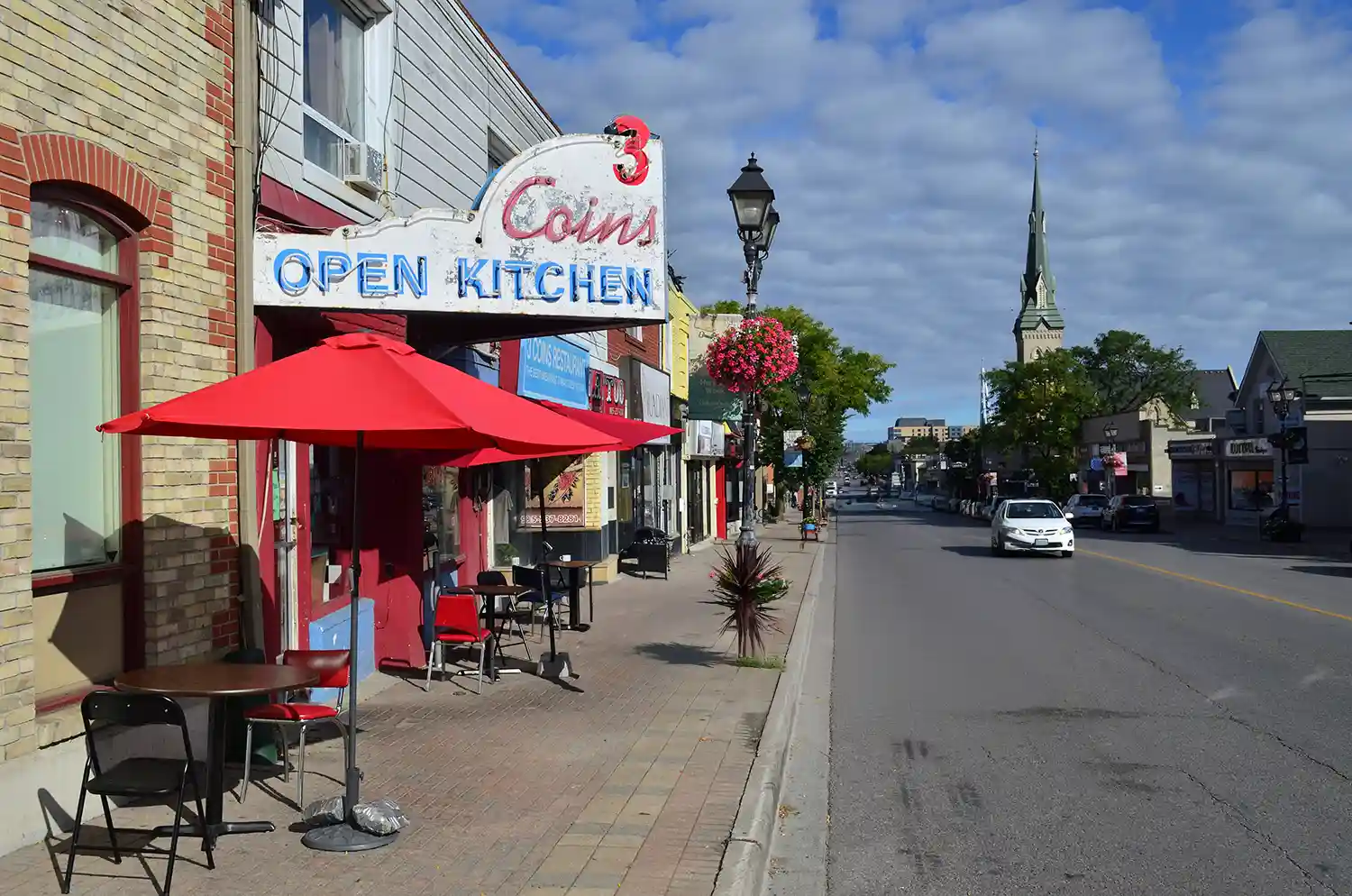 Street view of a small-town downtown with outdoor tables and a red umbrella in front of 3 Coins Open Kitchen restaurant, and a church steeple in the background.