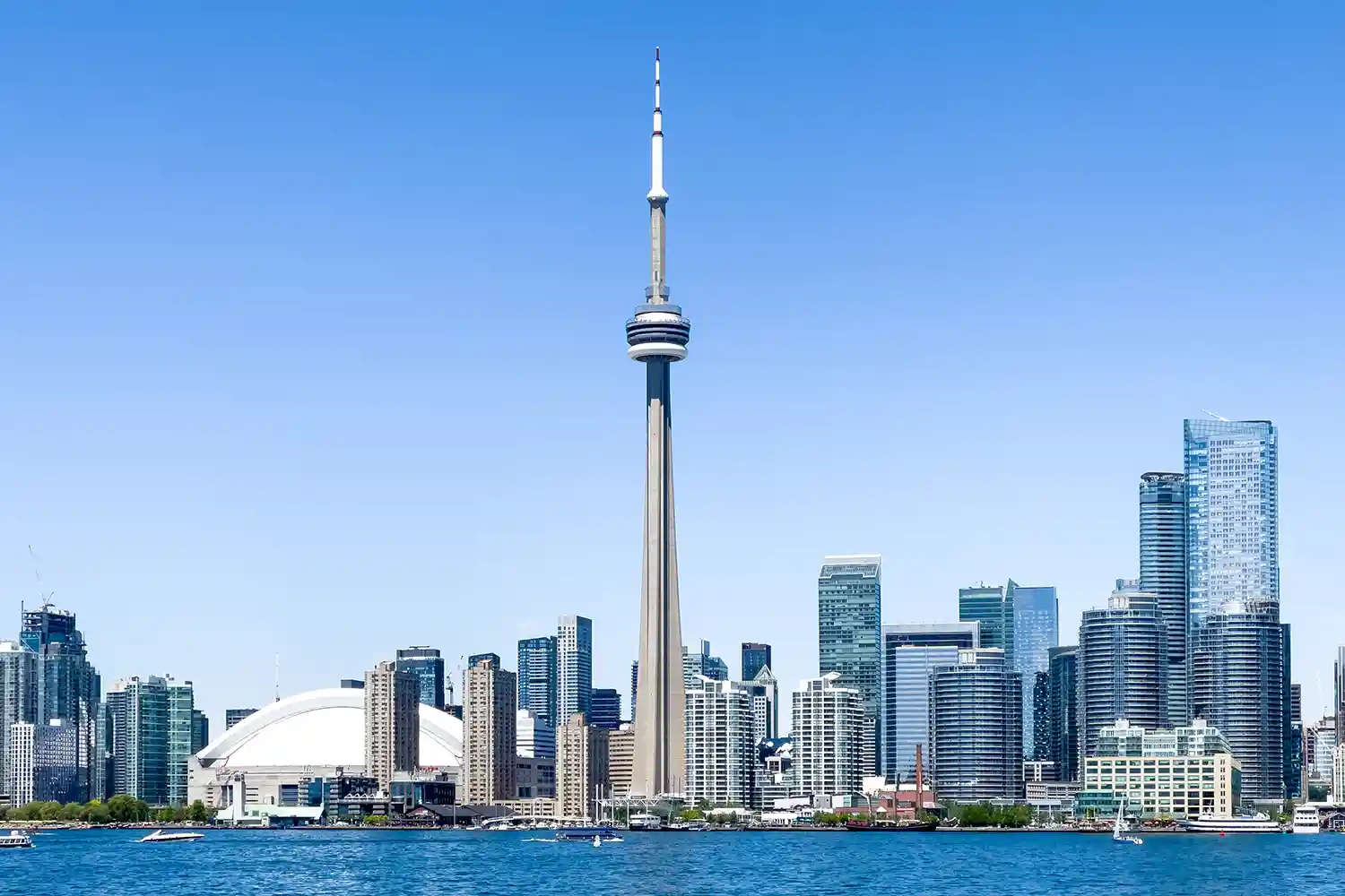 Toronto skyline with CN Tower prominently centered, surrounded by modern skyscrapers and Lake Ontario in the foreground under clear skies.