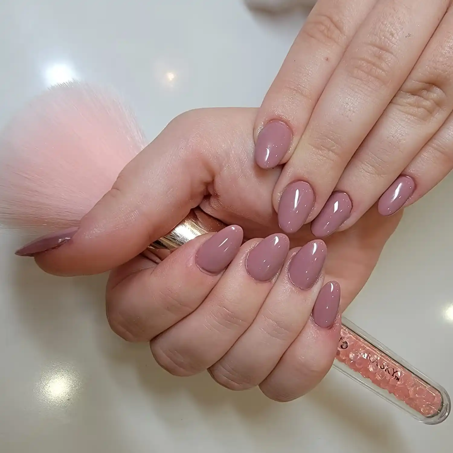 Hands with glossy mauve almond-shaped manicured nails holding a makeup brush with pink bristles and a clear handle filled with pink crystals.