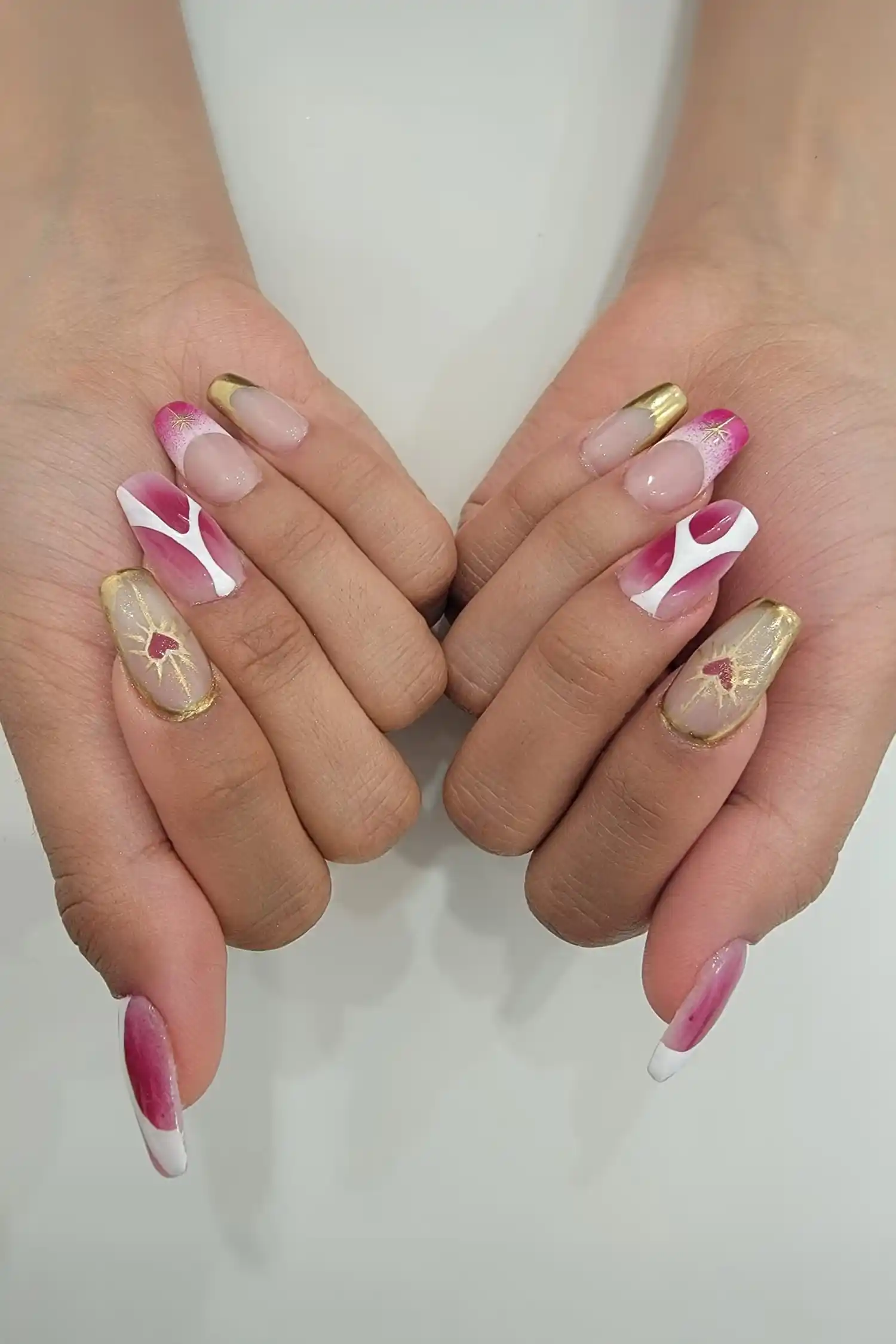 Hands with long nails decorated in pink, white, and gold designs including hearts and abstract patterns.