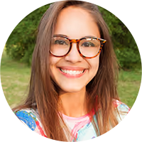 Smiling young woman with long brown hair and tortoiseshell glasses outdoors with green foliage background.