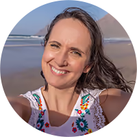 Smiling woman with dark hair wearing a white top with colorful embroidery takes a selfie outdoors near a body of water and mountains.