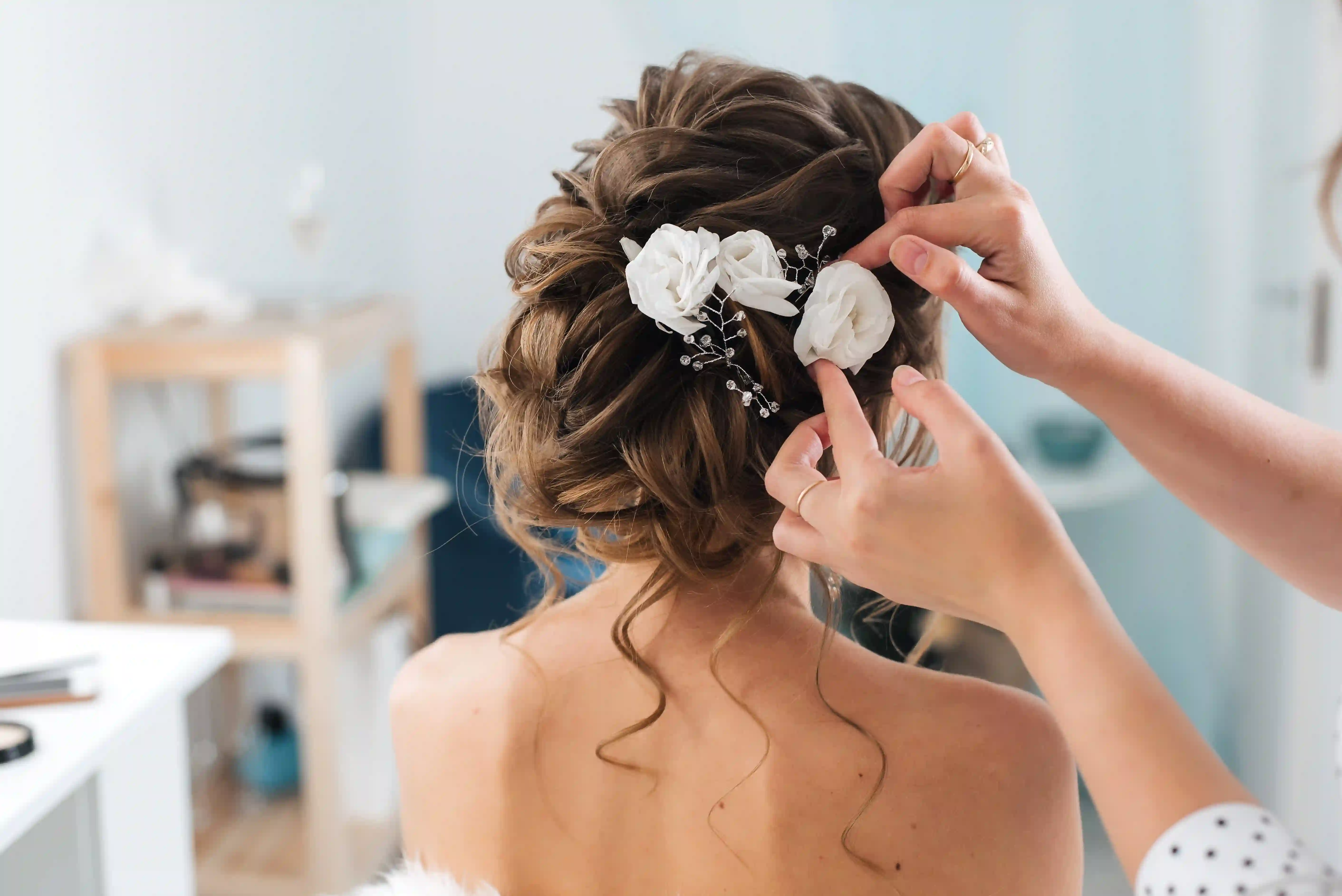Hands placing white flower decorations in a bride's curled updo hairstyle.