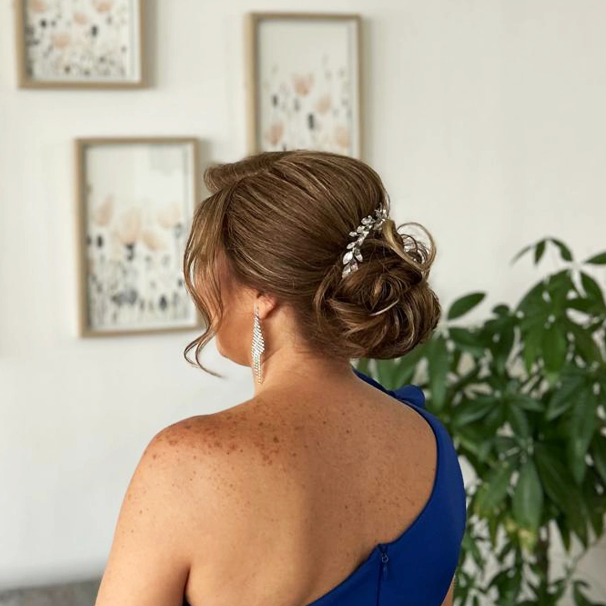Woman with light brown hair styled in an elegant low bun adorned with a silver leaf hair accessory, wearing a blue one-shoulder dress, viewed from the back.