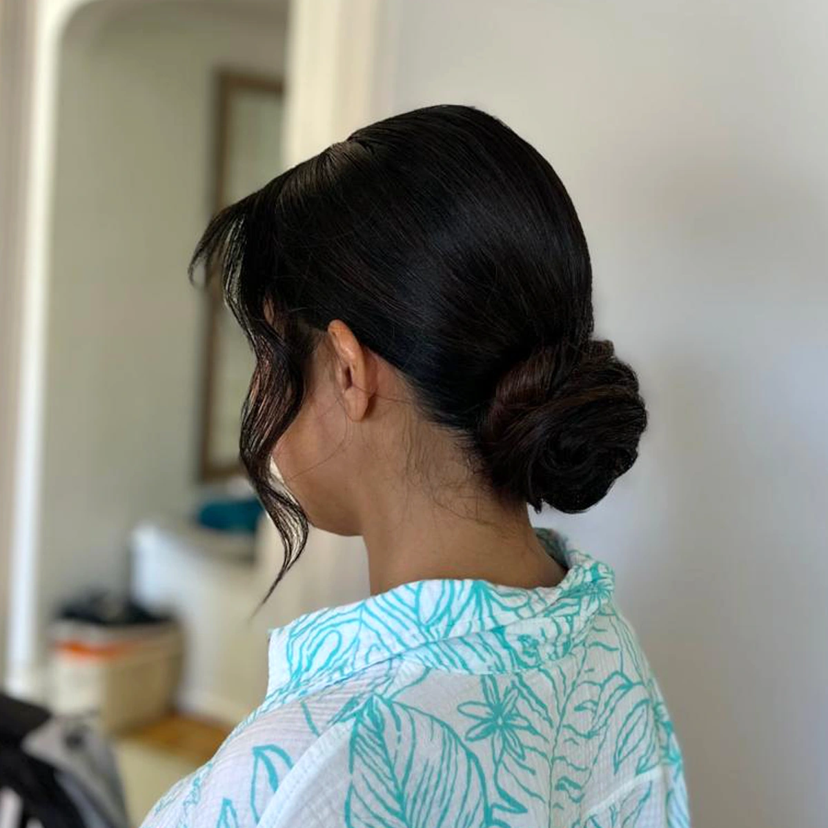 Woman with black hair styled in a low bun with a loose curl framing her face, wearing a white shirt with turquoise leaf patterns.