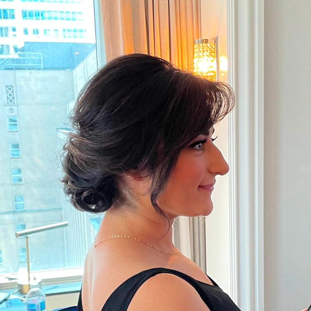 Woman with dark hair styled in an elegant low bun, wearing a black sleeveless top, standing indoors near a window with city buildings visible outside.