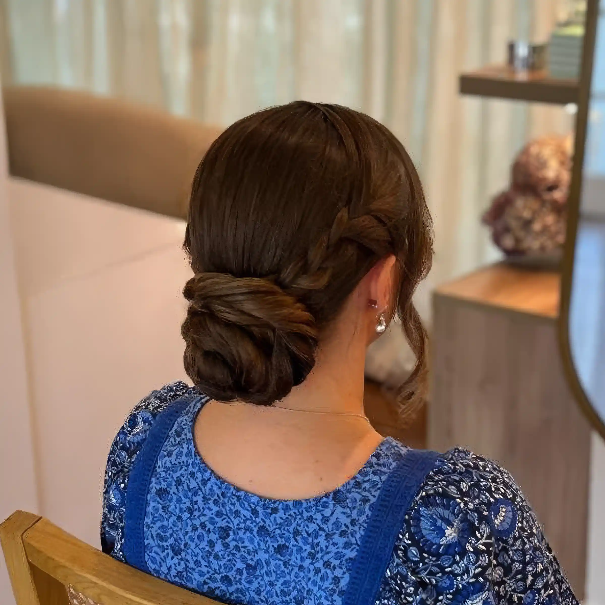 Back view of a woman with dark brown hair styled in a braided low bun wearing a blue floral dress.