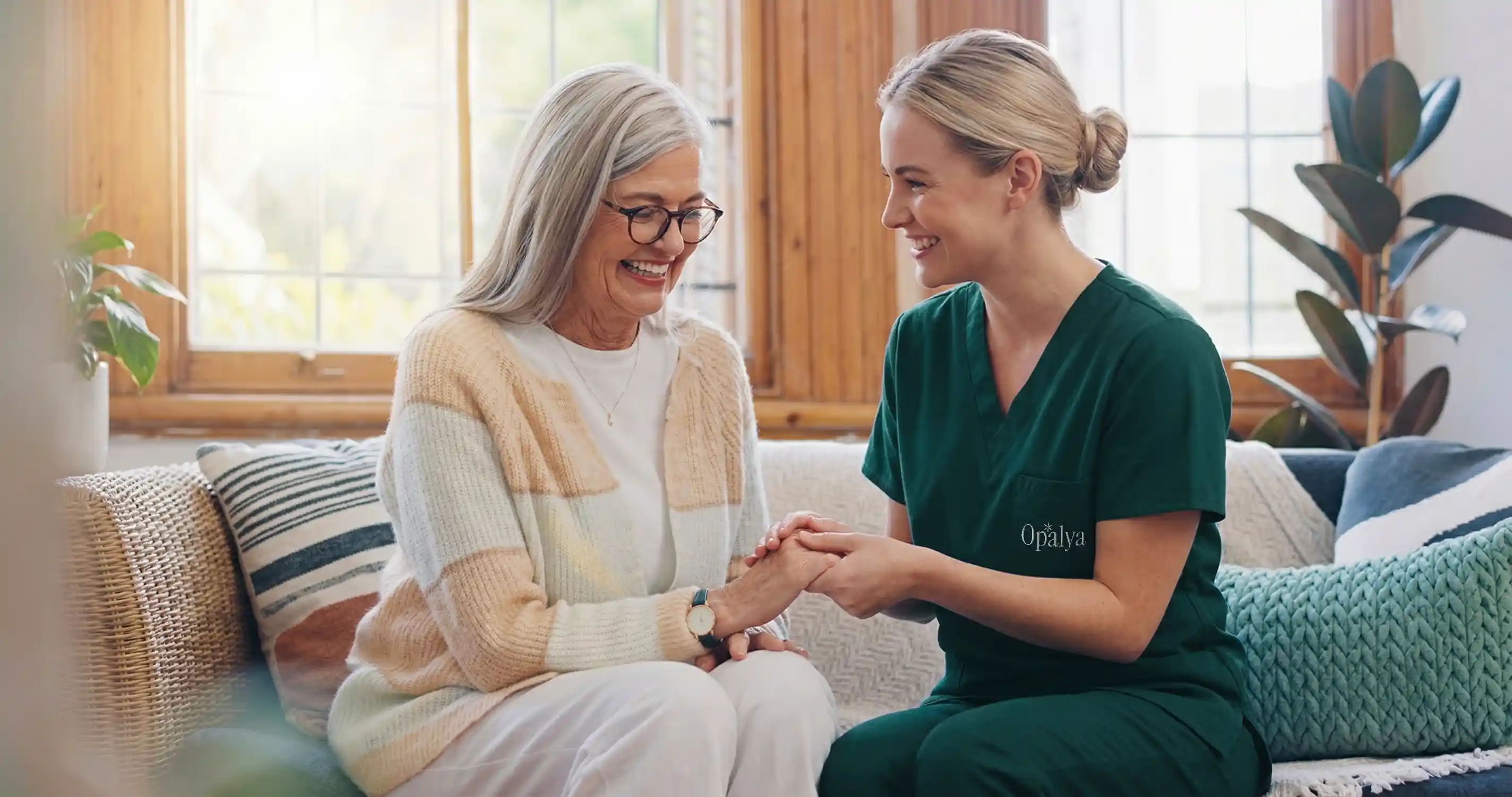 Smiling elderly woman sitting on a couch holding hands with a cheerful female caregiver in green scrubs.