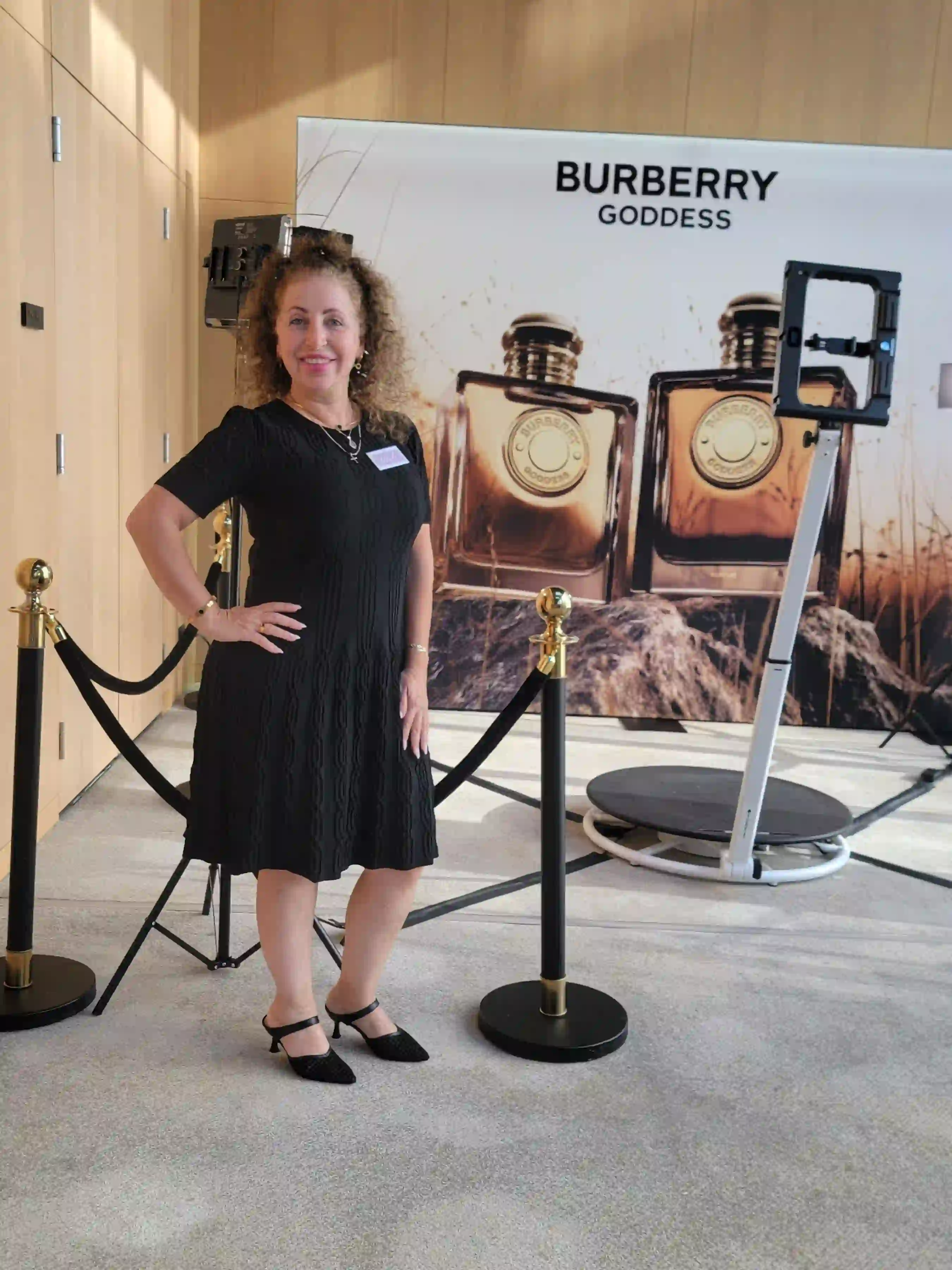Woman in a black dress posing near a Burberry Goddess perfume advertisement with two large perfume bottles displayed.