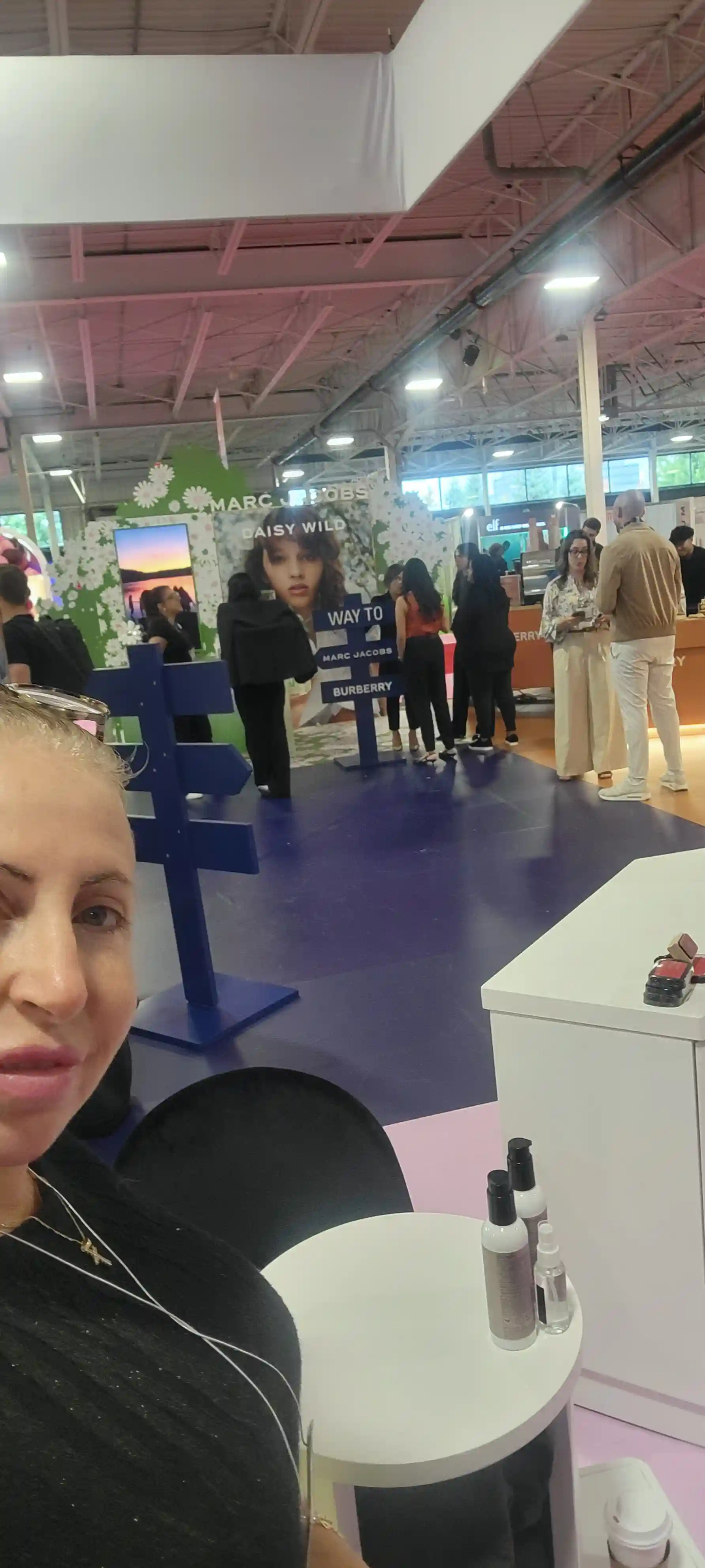 Woman taking a selfie at an indoor event with booths and people, including a Marc Jacobs display in the background.