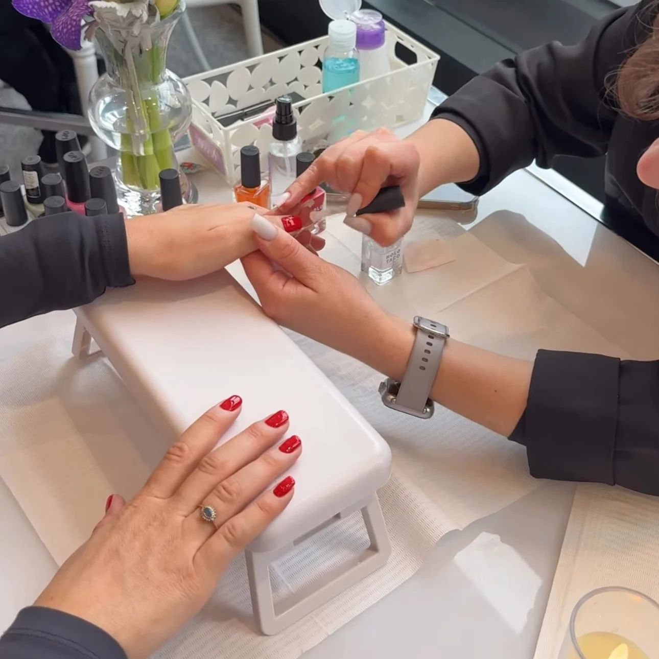 Manicurist painting red nail polish on a customer's nails at a nail salon with nail care products nearby.