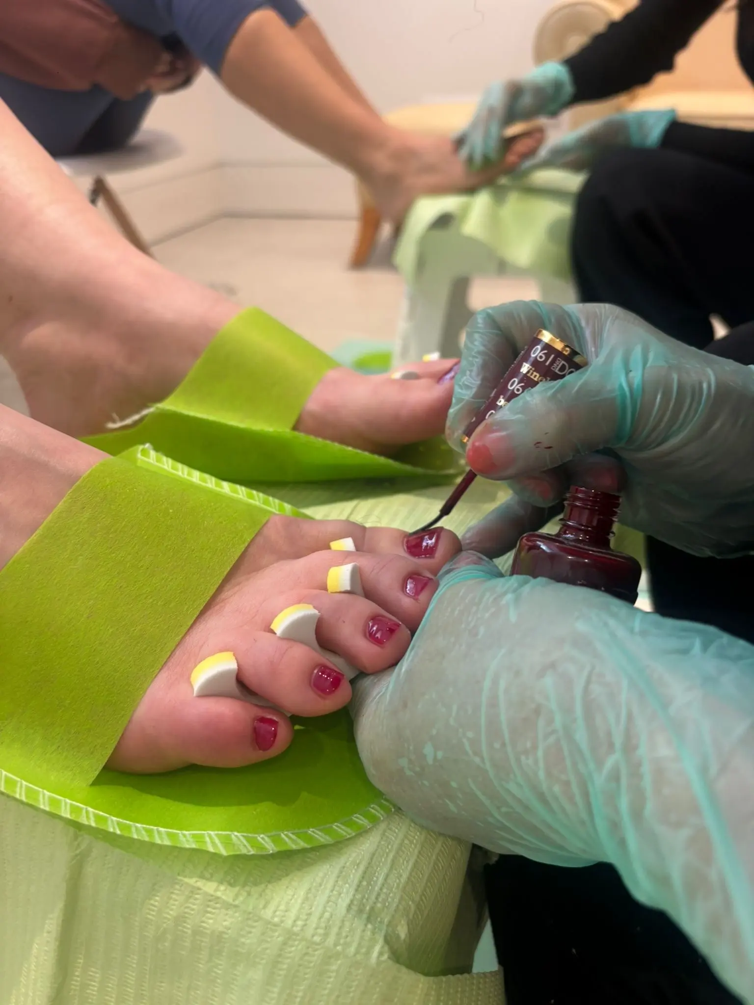 Close-up of a pedicure in progress with a technician applying dark red nail polish to toenails separated by white and yellow toe separators.