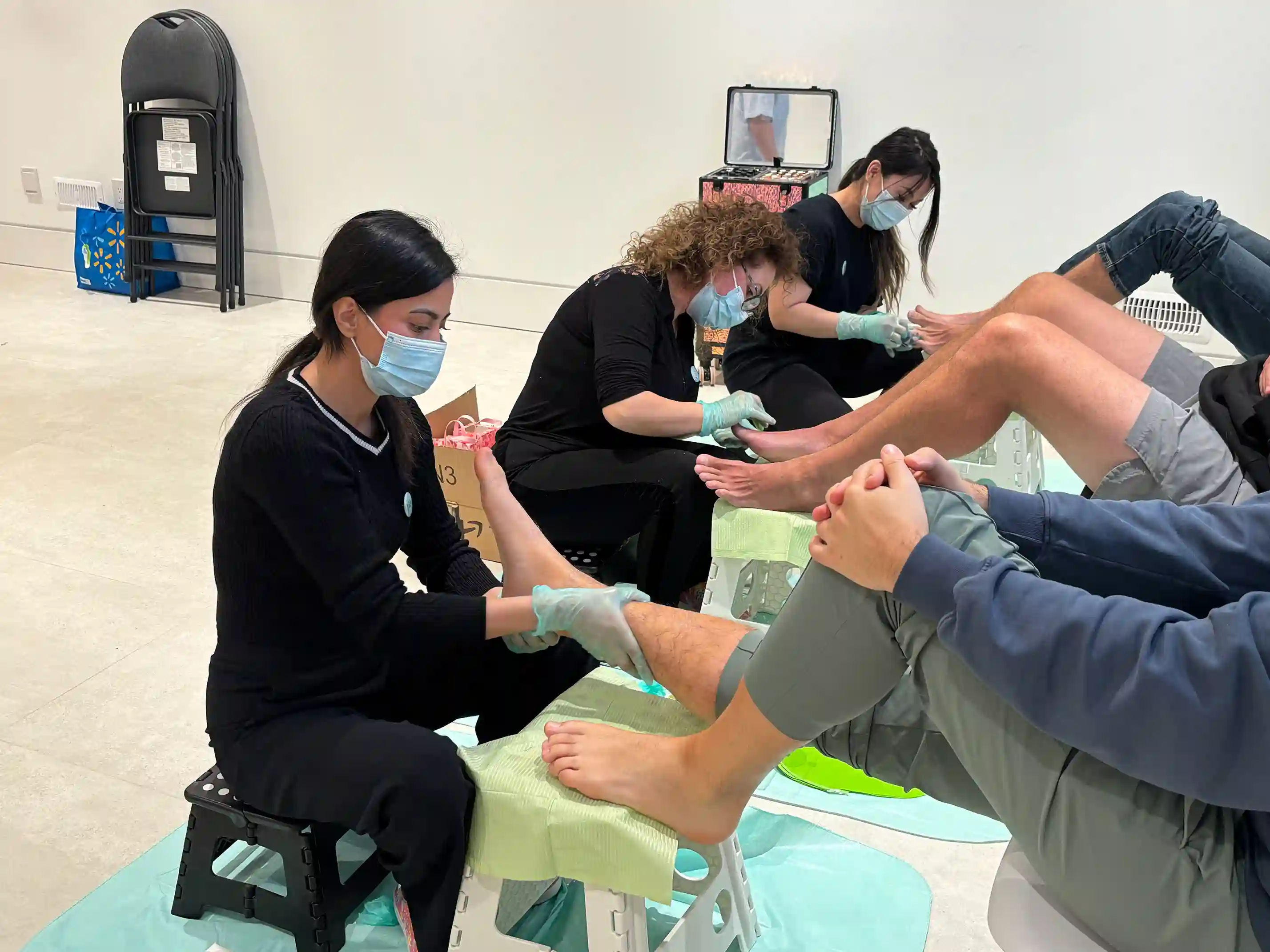Three women wearing masks and gloves giving pedicures to three seated men in a clean, indoor setting.