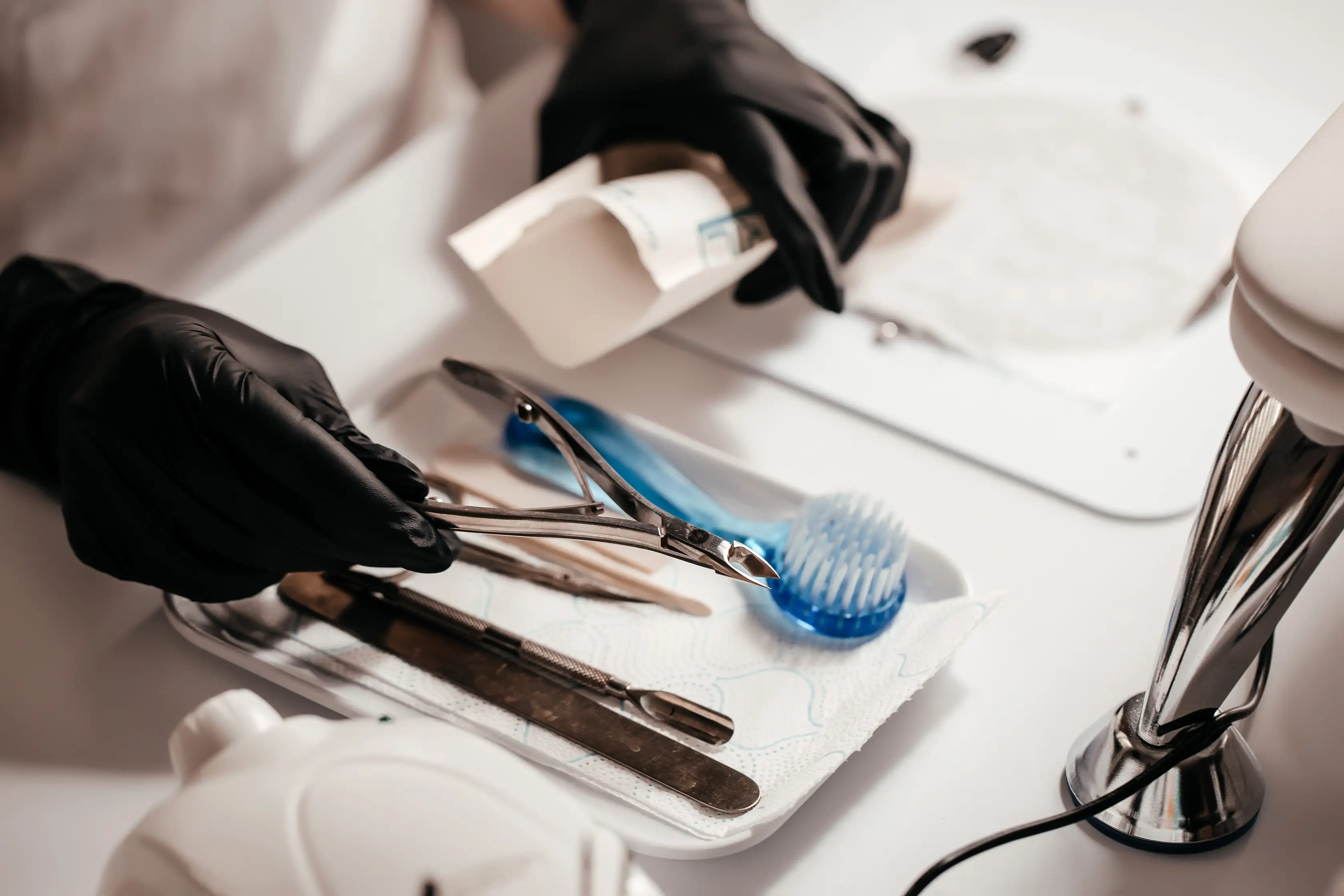 Hands in black gloves holding sterilized nail clippers over a tray with manicure tools and brush.