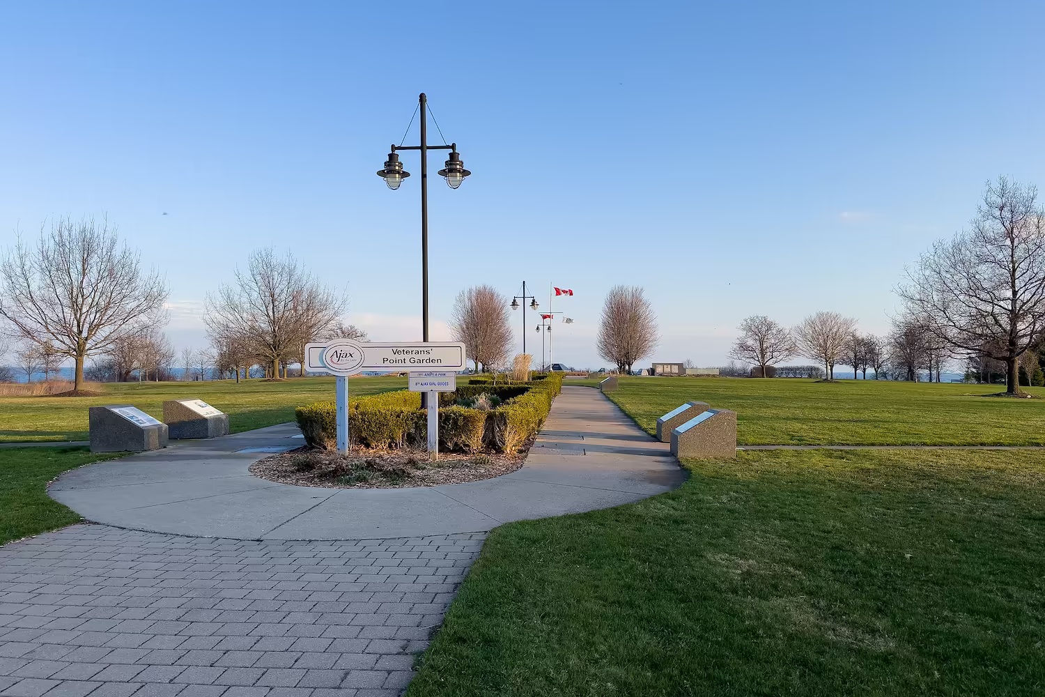 Pathway through Veterans' Point Garden with signs, leafless trees, grass, and flagpoles in Ajax, Canada.