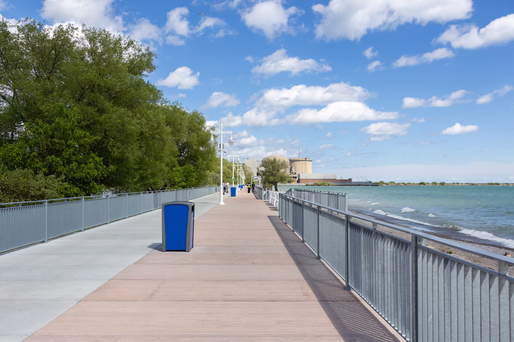 Boardwalk along a waterfront with trees on left and railing on right under blue sky with clouds.
