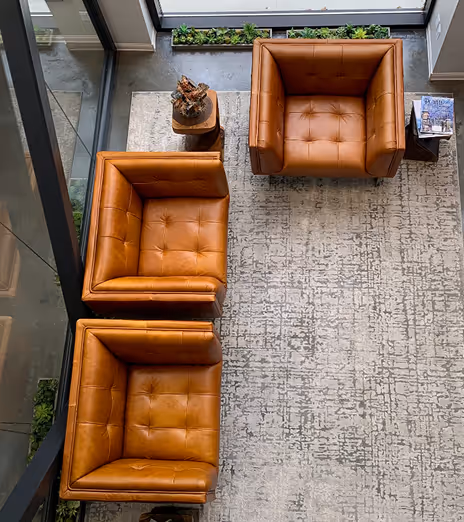 Overhead view of three tan leather armchairs arranged on a textured light gray rug near large windows, with small wooden side tables holding magazines and a decorative item.