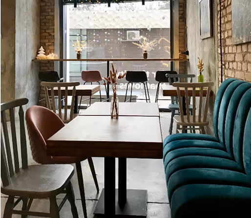 Cozy cafe interior with wooden tables, mismatched chairs, and a green velvet bench near a window decorated with hanging lights and small plants.