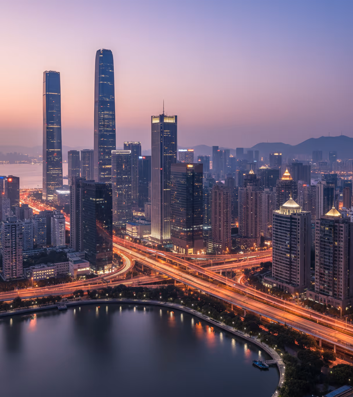 Cityscape at dusk with illuminated skyscrapers, highways, and a curved waterfront reflecting city lights.