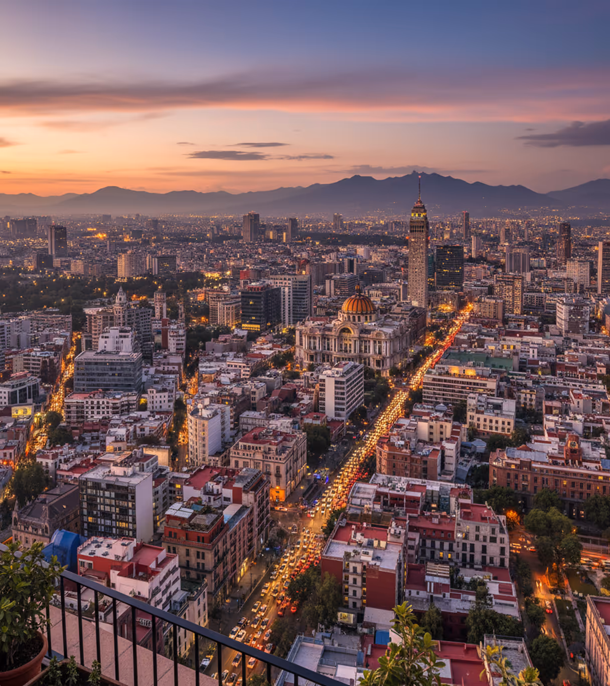 Aerial view of Mexico City at sunset with the Palacio de Bellas Artes and Torre Latinoamericana tower amidst city buildings and mountain backdrop.
