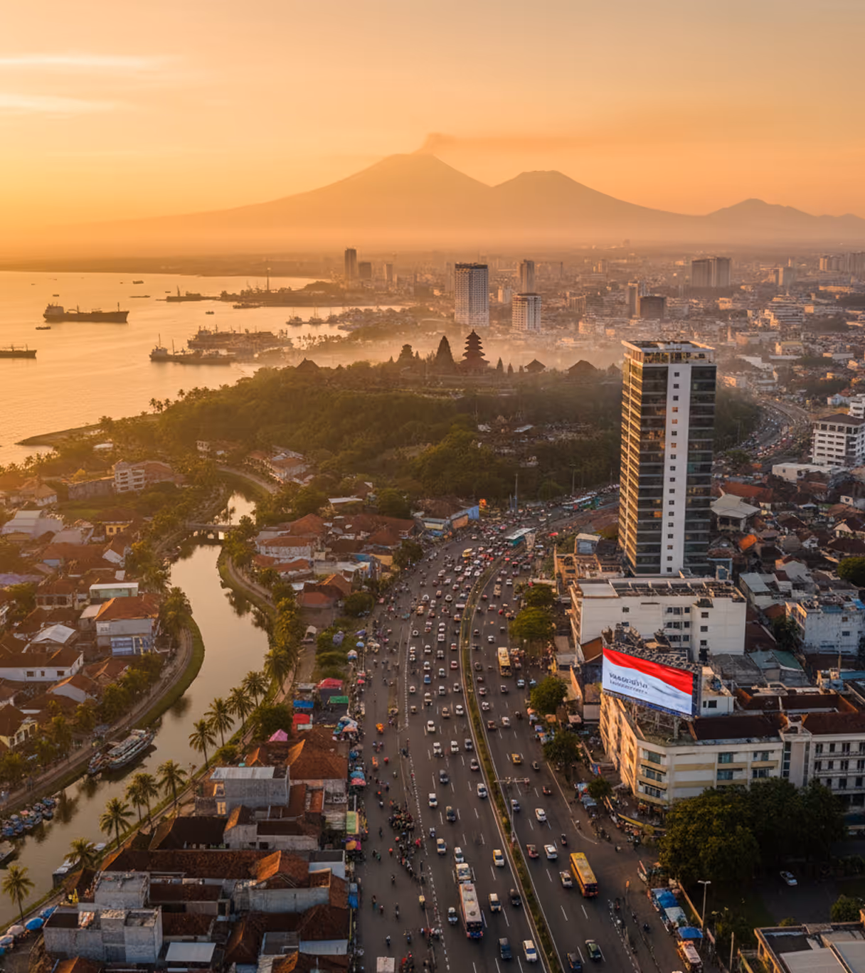 Aerial view of a busy urban highway next to a canal, dense buildings, a green park with a pagoda, large ships in the harbor, and mountains in the distant sunset haze.